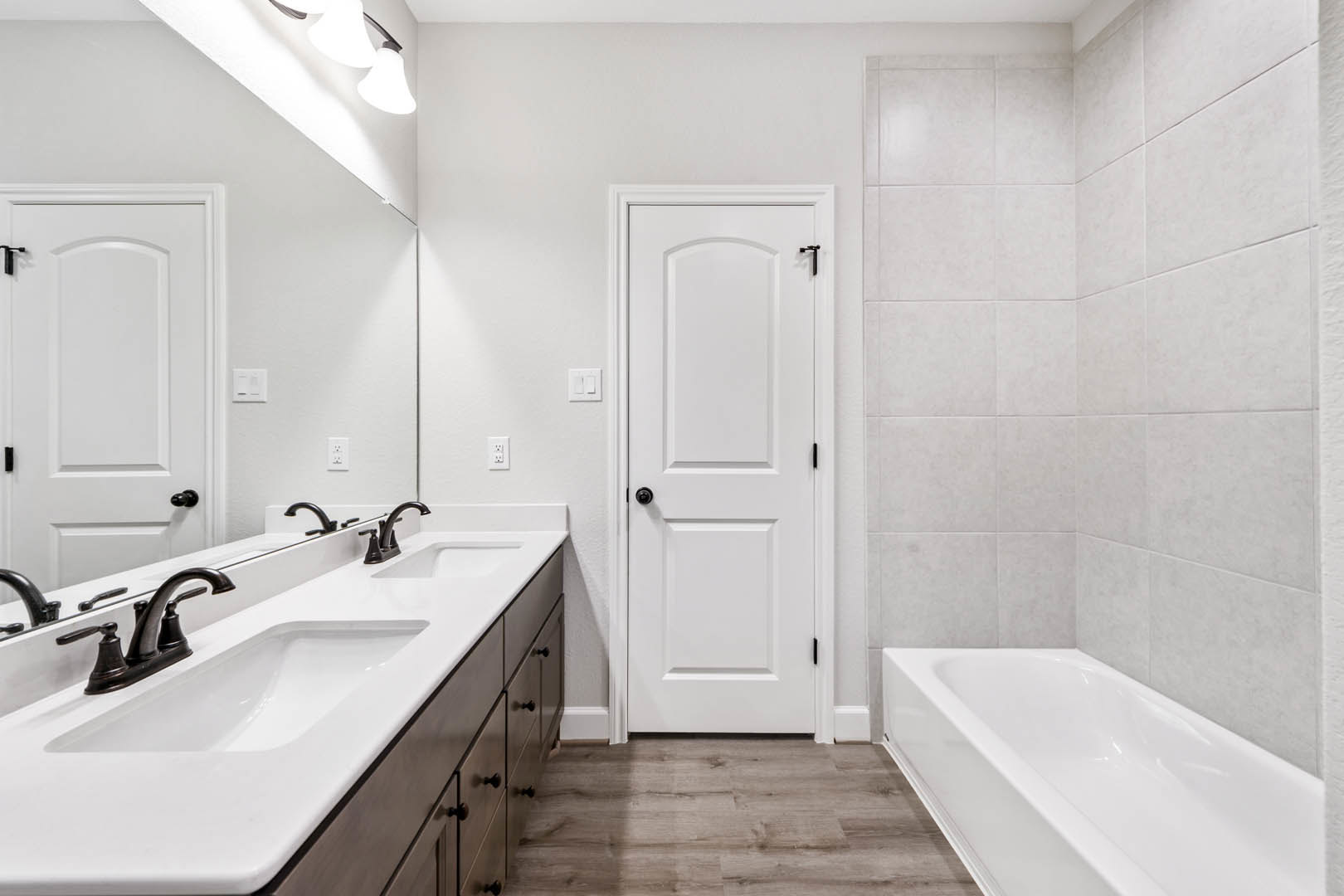 White bathroom door with black handles, white sink with chrome faucet, light tile walls, and partial view of a white bathtub.