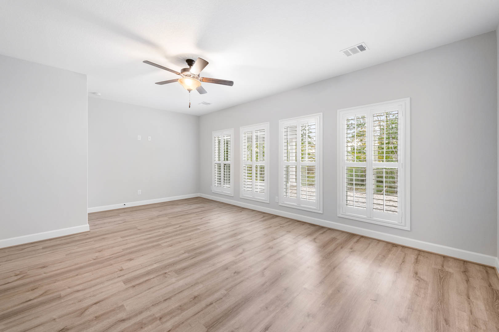 Living room with wood laminate flooring, white walls, ceiling fan with light, multiple windows fitted with white blinds, and a square white air vent.