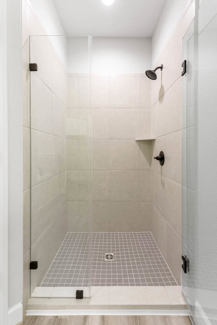 Walk-in shower with glass door, wall-mounted shower head, white tile walls, black tile border, and floor drain