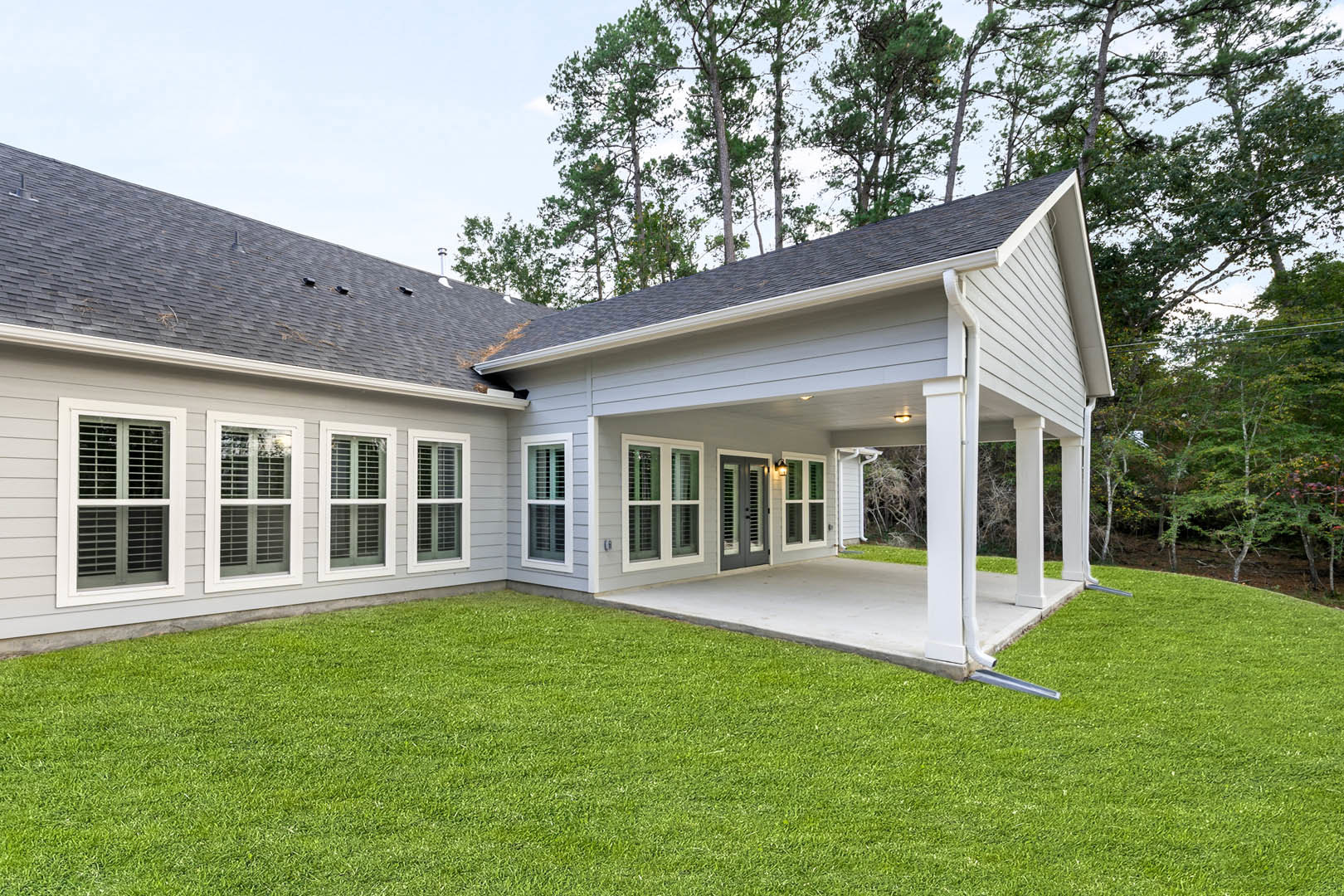 Modern house with white-framed windows, covered patio featuring wood posts, and green lawn bordered by landscaping plants