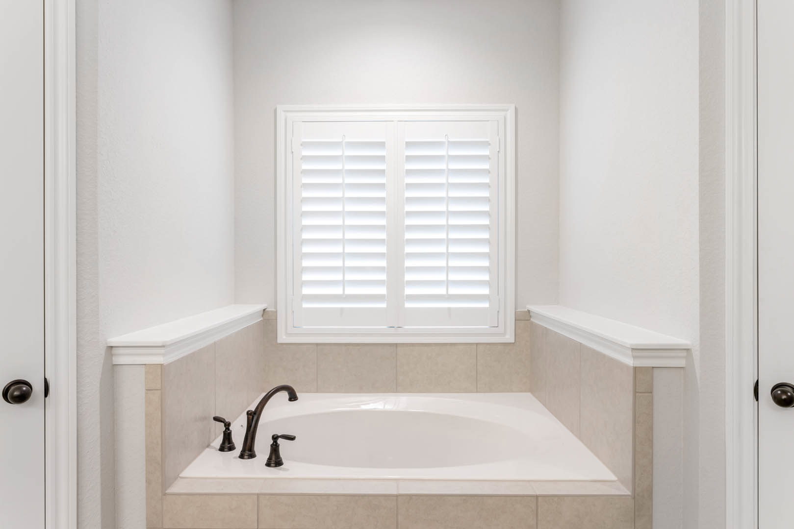 Freestanding white bathtub beneath a window with white blinds, chrome faucet, light gray tile walls, and black decorative accent on tub ledge