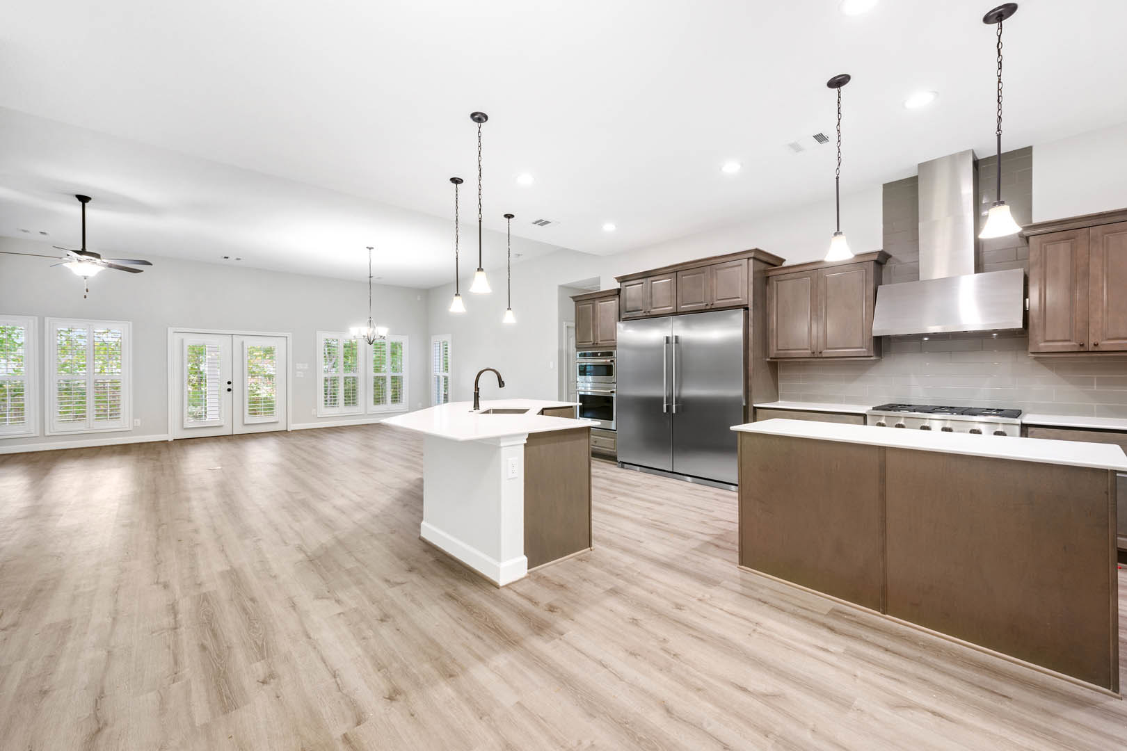 Kitchen with wood flooring, white cabinetry, stainless steel refrigerator with double handles, central island topped with light fixture, white double doors with blinds, chain