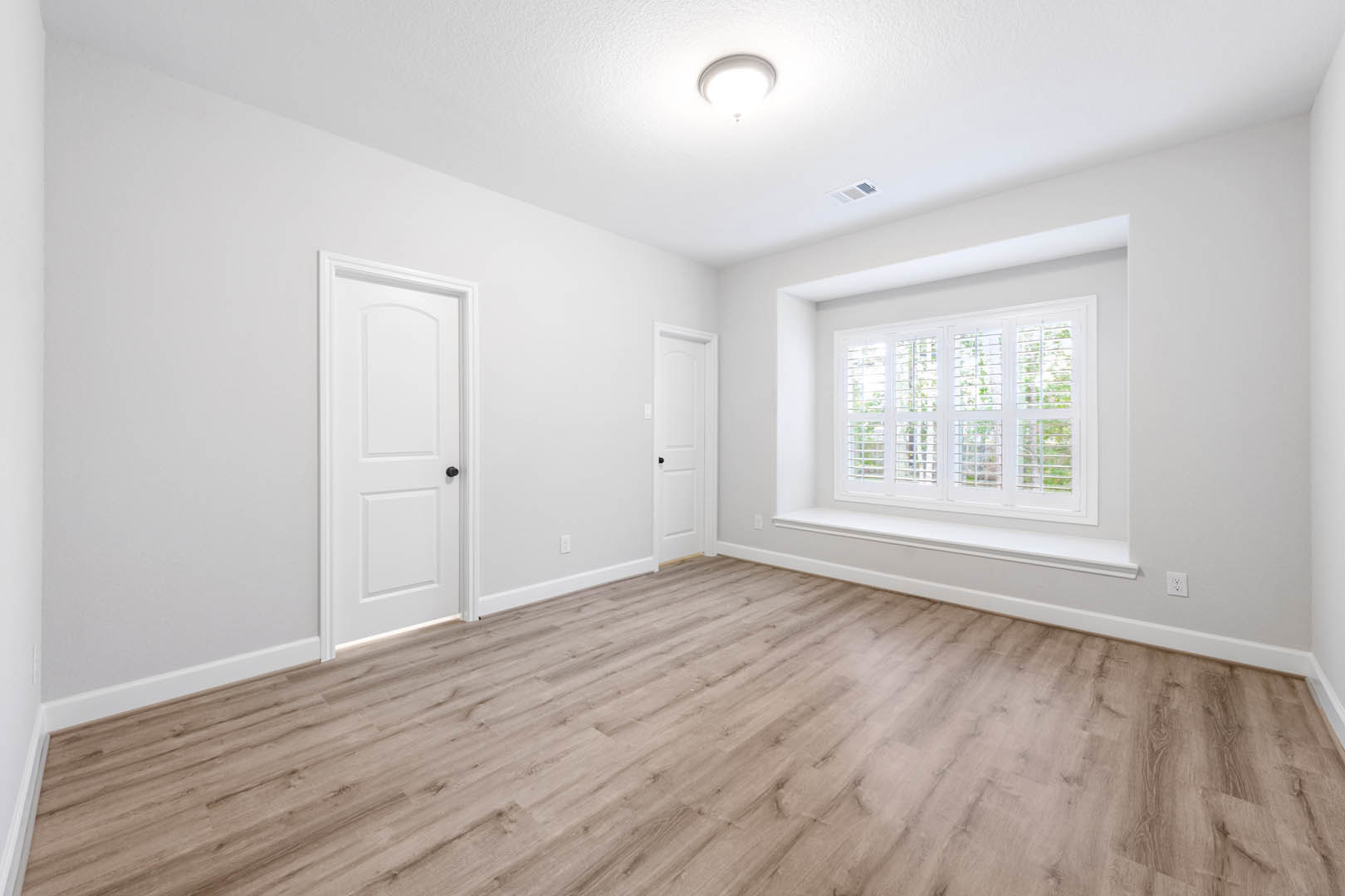 Wood flooring in a bright room with white walls, white door with black knob, window with blinds, and ceiling light fixture