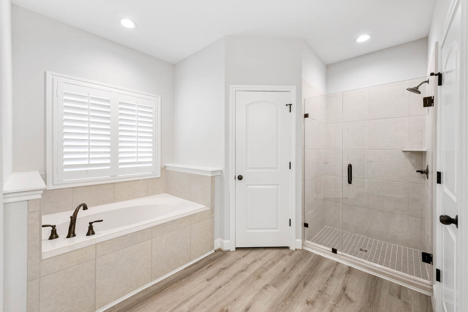 Bathroom with freestanding white bathtub, glass-enclosed tile shower, wood flooring, white door with black hardware, window with white shutters