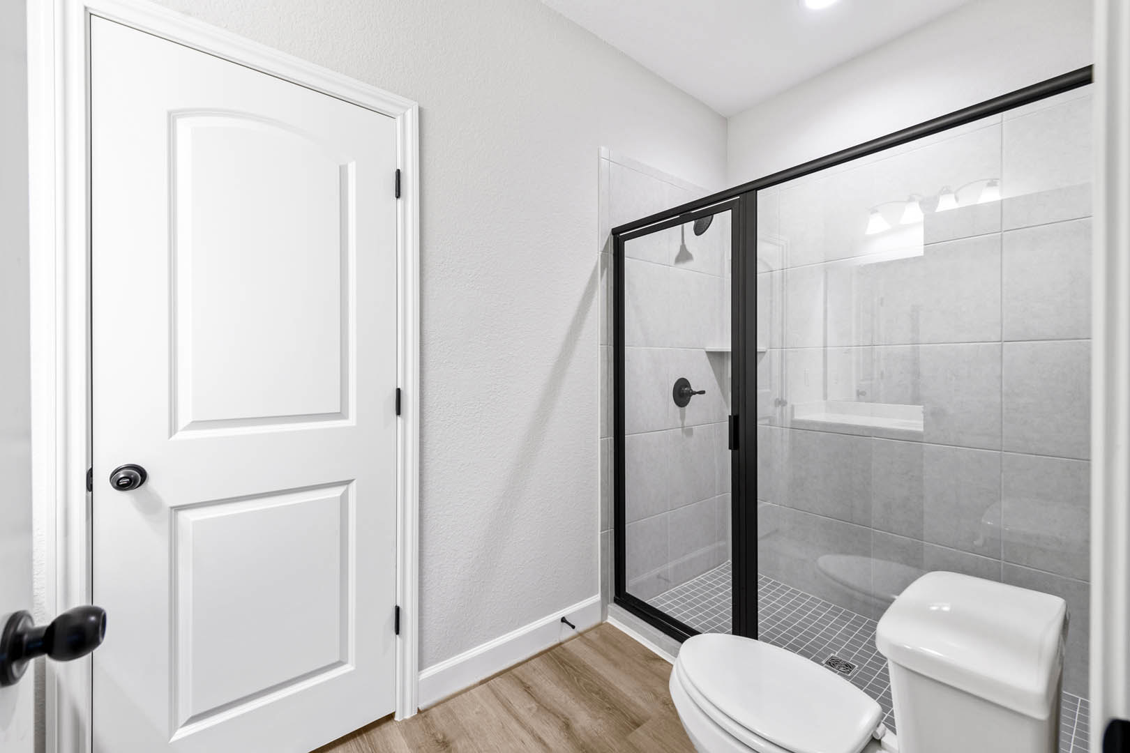 Bathroom with glass shower door, white toilet, grey tile wall, and white door with black hardware
