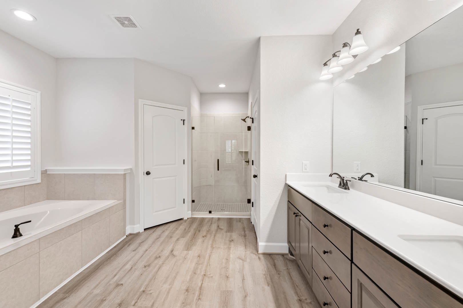Bathroom with wood plank flooring, white walls, double sink vanity, black hardware on white door, glass shower enclosure with built-in shelf, chrome faucet, window with white