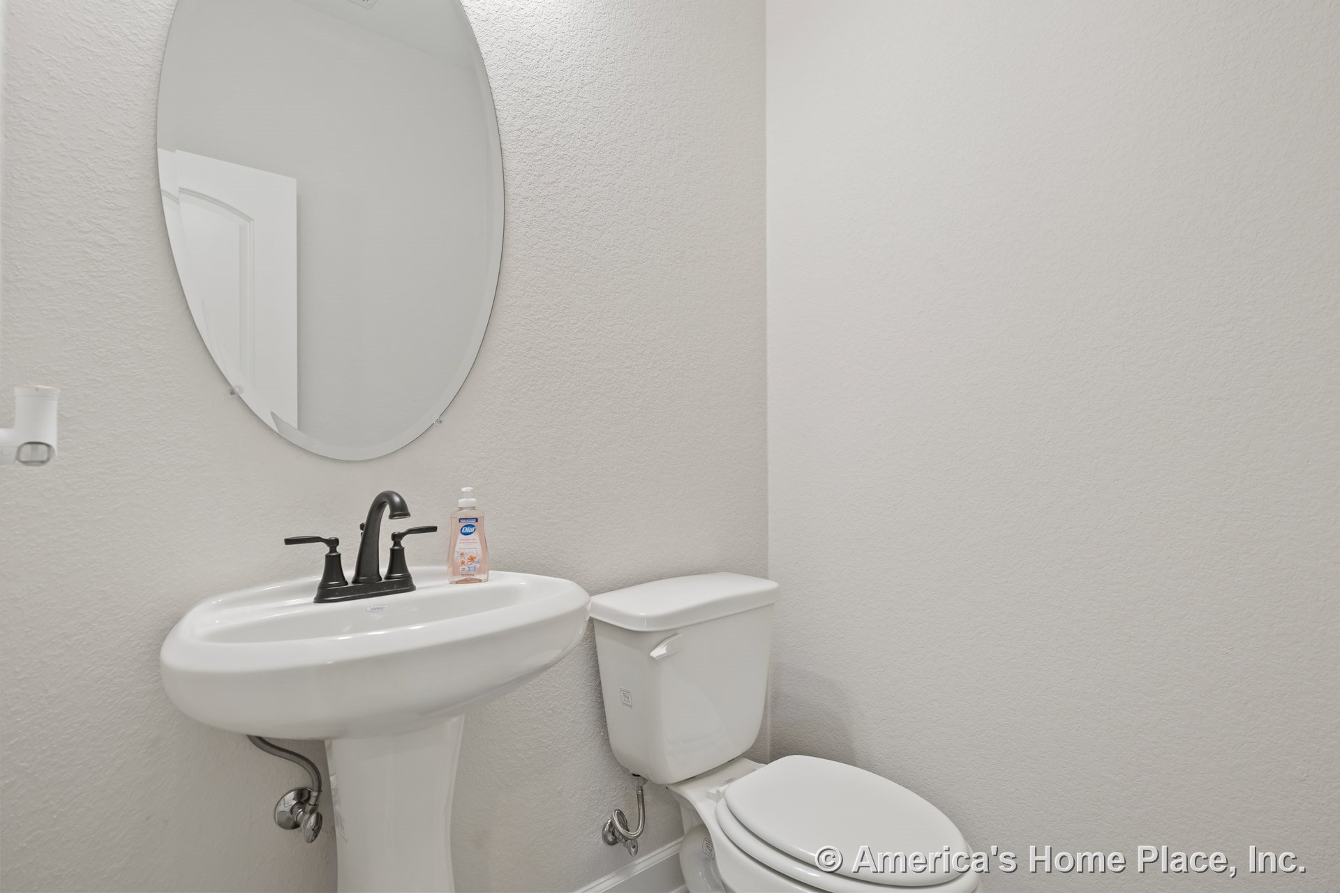 Pedestal sink with black faucet beneath an oval wall mirror, white toilet adjacent, textured light-colored walls, minimal trim, compact powder room layout.