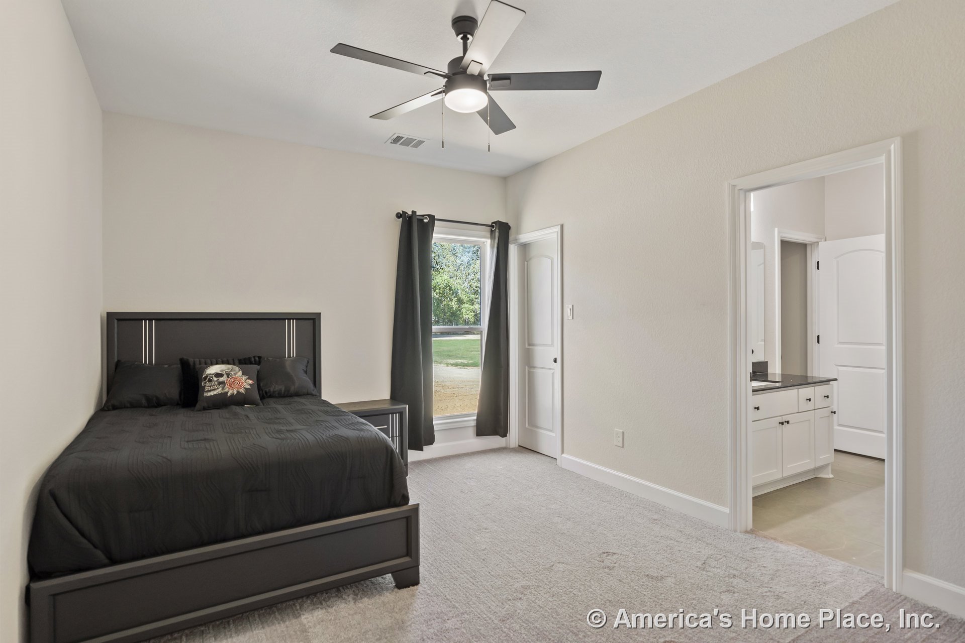 Carpeted bedroom with large window framed by black curtains, ceiling fan and light fixture overhead, white trim and baseboards, adjoining bathroom featuring white cabinetry and