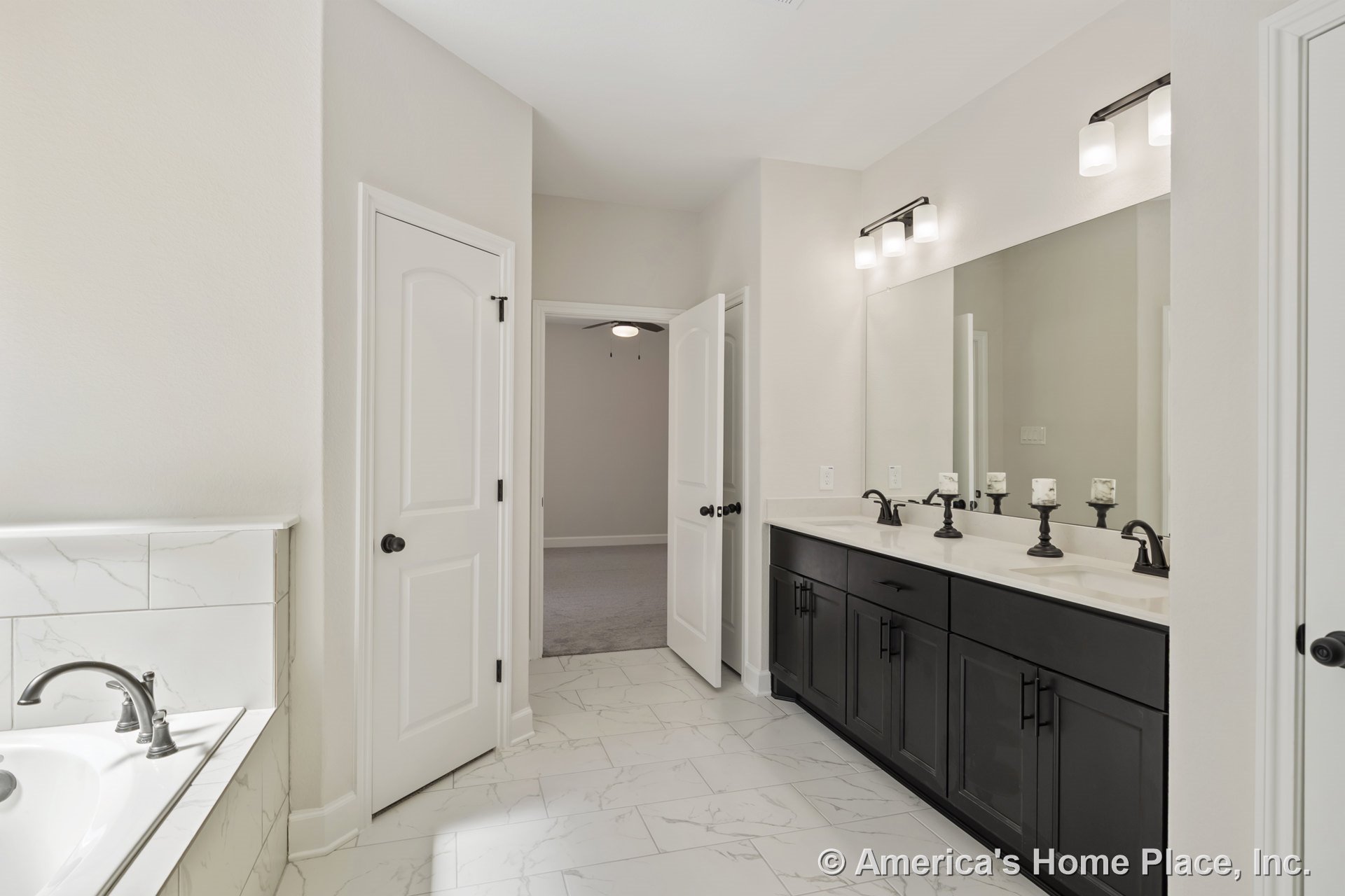 Double vanity with dark wood cabinets and sleek faucets, marble-look tile flooring, built-in bathtub, large wall mirror, modern overhead lighting, white walls, and door trim.