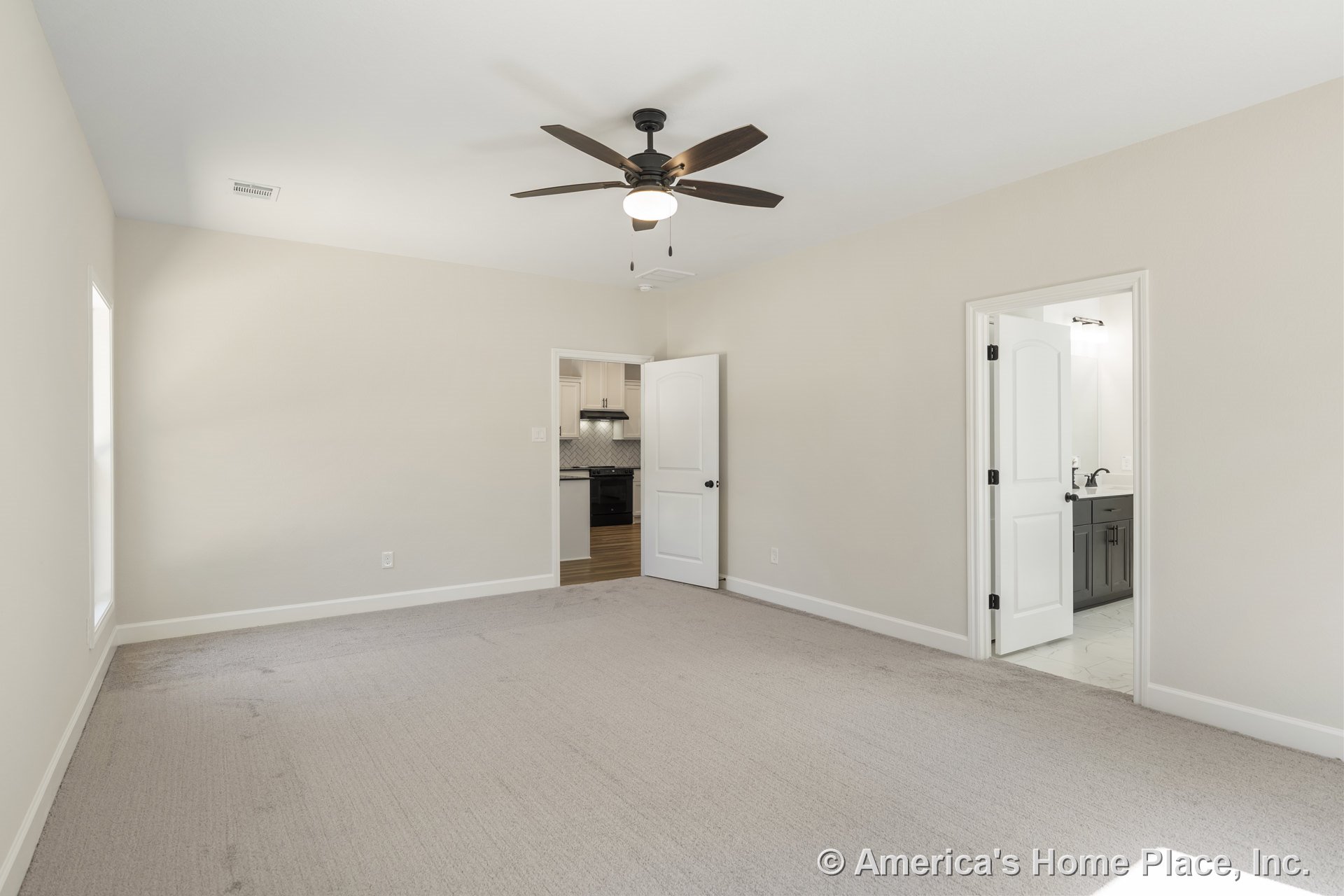 Spacious bedroom with light carpet flooring, ceiling fan and light fixture, white trim and baseboards, paneled interior doors, bathroom vanity with dark cabinetry, and kitchen