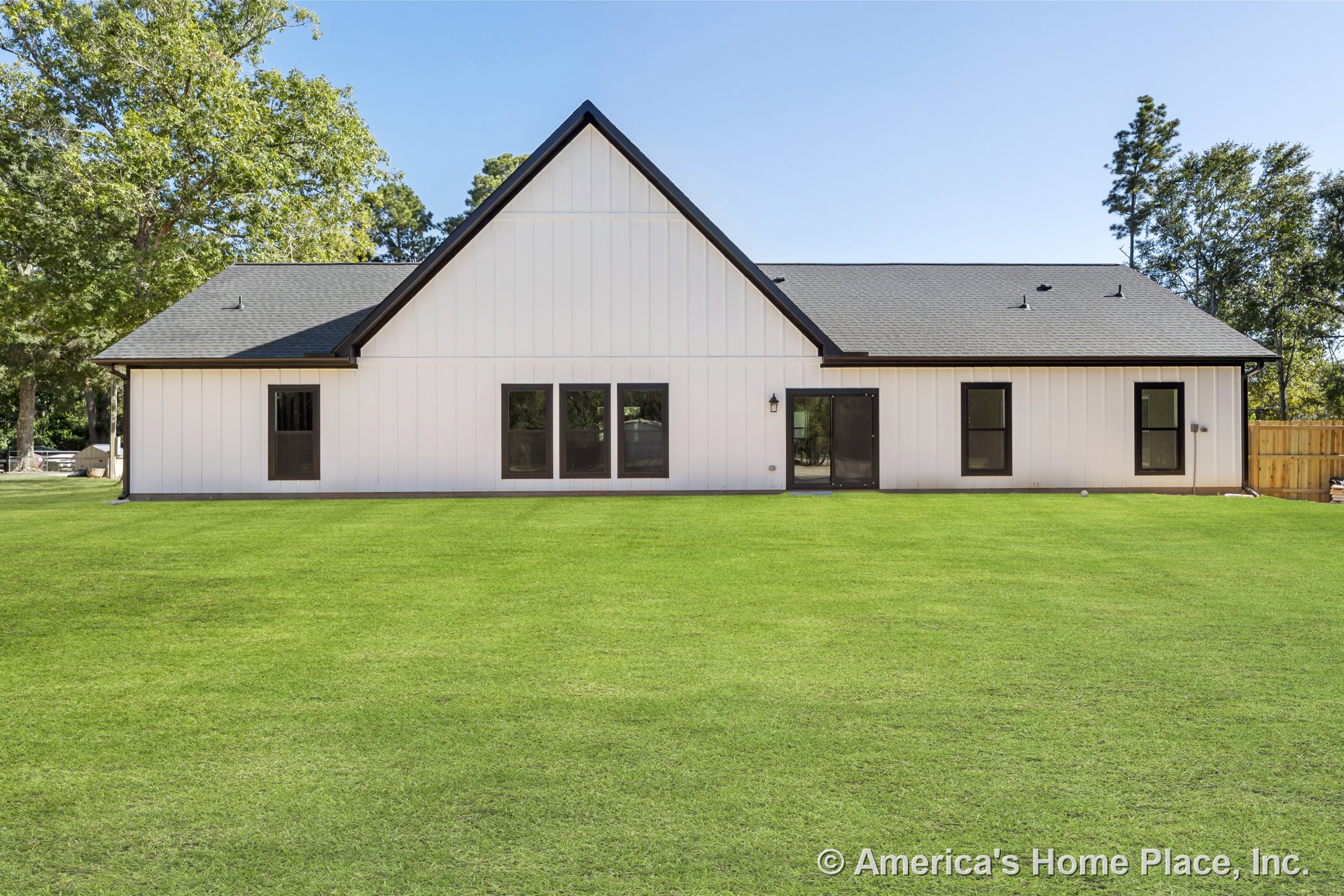 Vertical board and batten siding with black trim, multiple large windows, gable roof, glass-paneled exterior door, outdoor wall light fixture, manicured lawn, and partial fence.