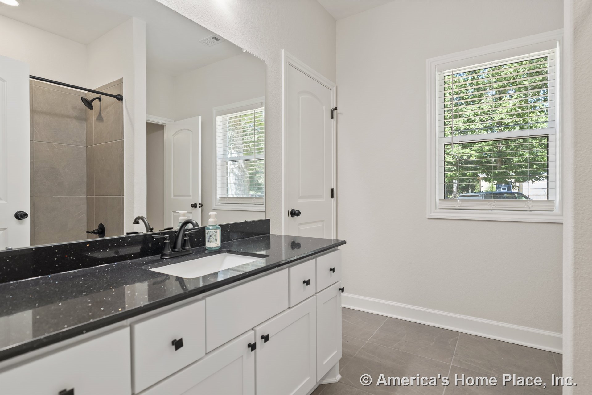 Black solid surface vanity with white shaker cabinets, large wall mirror, tiled walk-in shower, gray tile flooring, window with blinds, matte black fixtures, and white door trim.