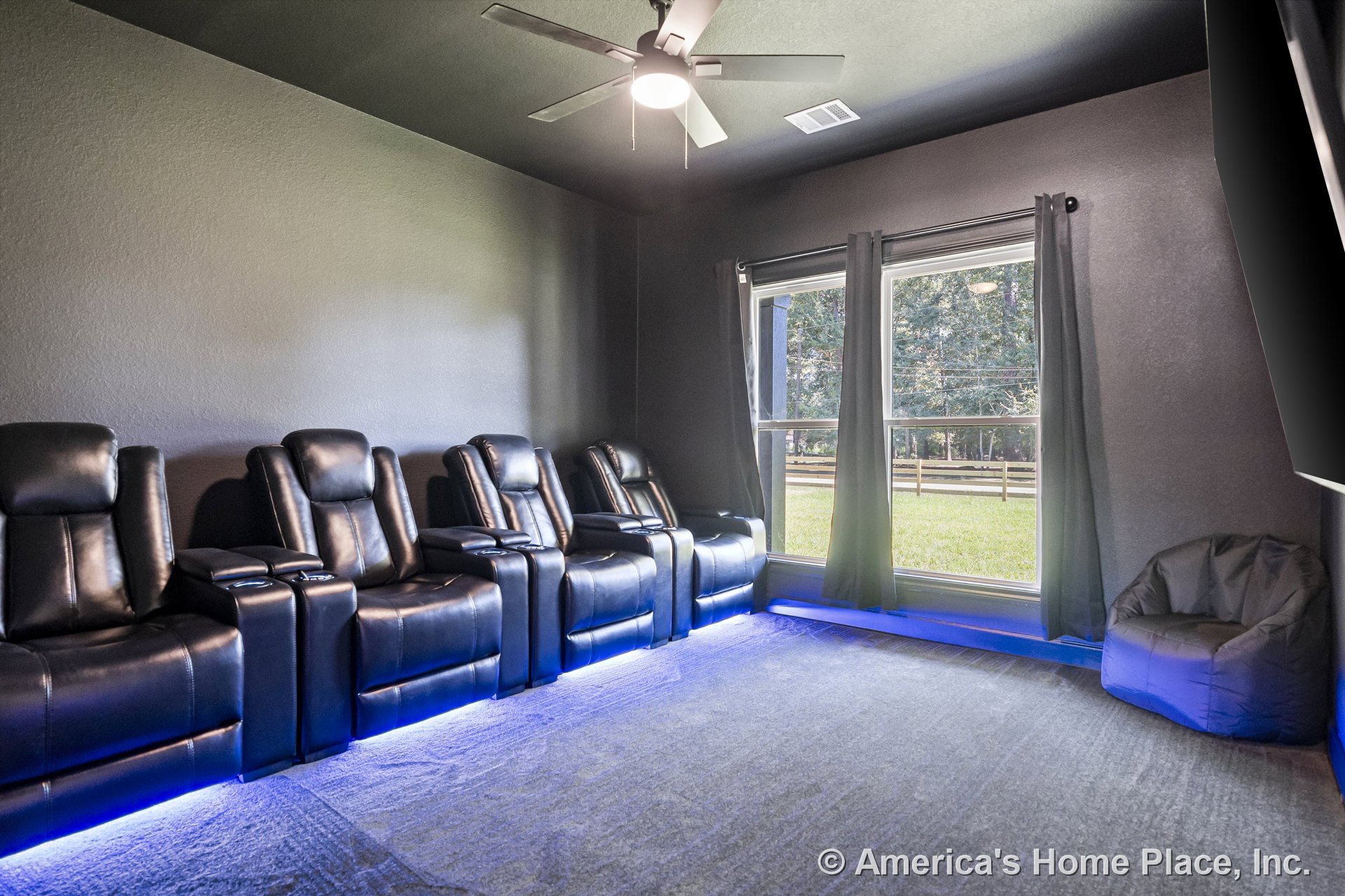 Home theater room with wall-to-wall carpet flooring, dark painted walls, large double windows framed by built-in blackout curtains, recessed LED accent lighting, and ceiling fan
