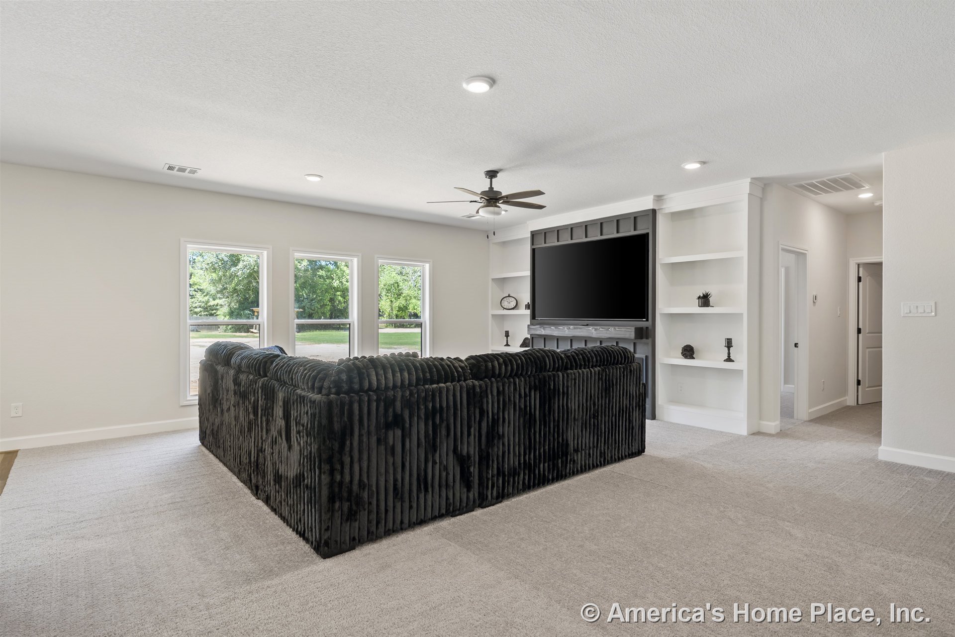 Built-in shelving flanks a wall-mounted television, three tall windows brighten the carpeted floor, ceiling fan with lights hangs overhead, white trim and baseboards accent neutral