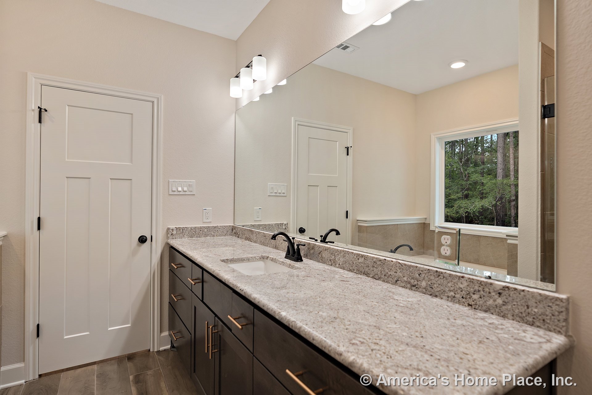 Granite countertop with dual sinks and matte black faucets atop dark wood cabinetry, large wall mirror spanning the vanity, recessed ceiling light, window with outdoor view, tile
