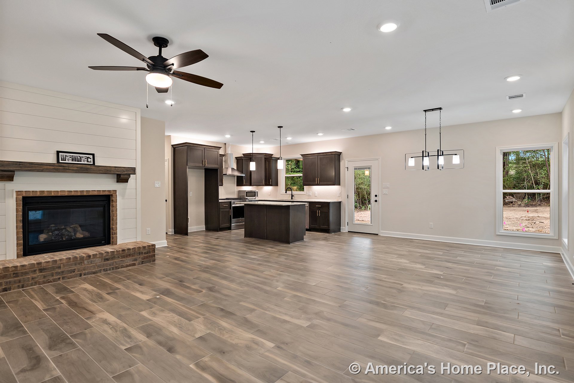Open concept living area with wood-look tile flooring, brick fireplace framed by shiplap trim, modern kitchen featuring dark cabinetry and pendant lights above the island, large