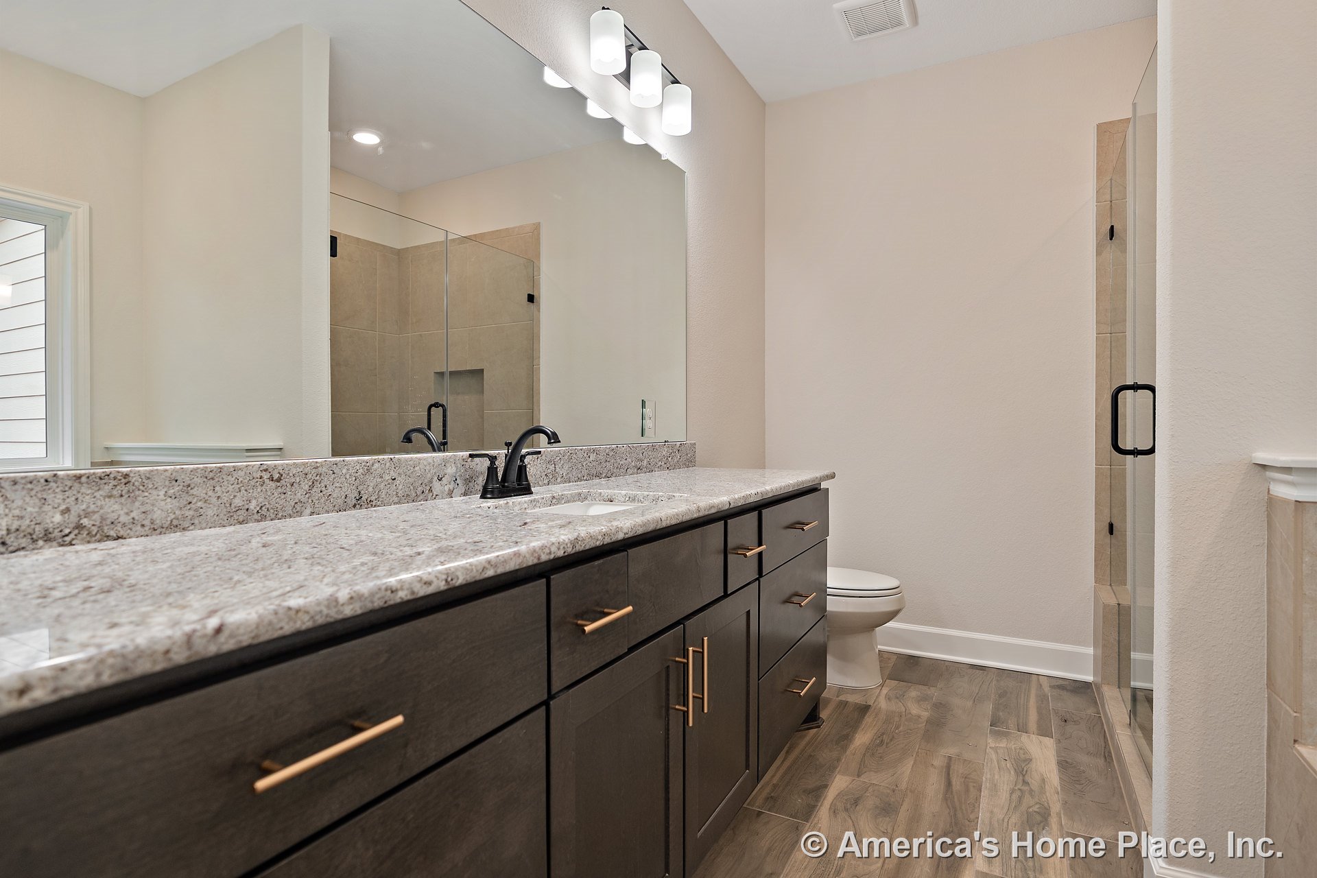 Double vanity with granite countertops and dark wood cabinetry, glass-enclosed shower with tile flooring, large wall mirror, wall-mounted vanity lights, modern faucets, and