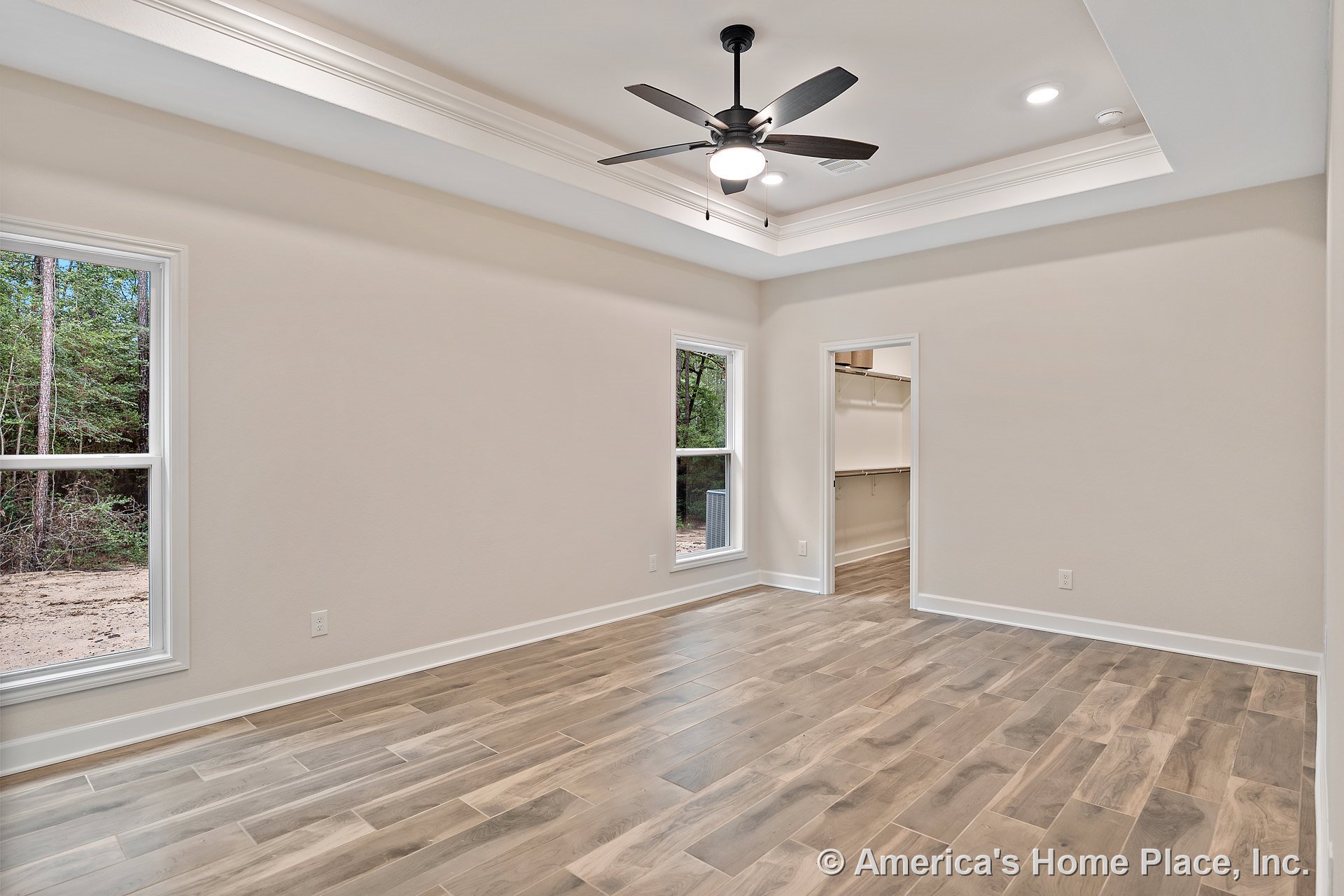 Bedroom with wood-look tile flooring, tray ceiling featuring recessed lights and ceiling fan, two large windows, walk-in closet entry, neutral walls, baseboards, and door trim.