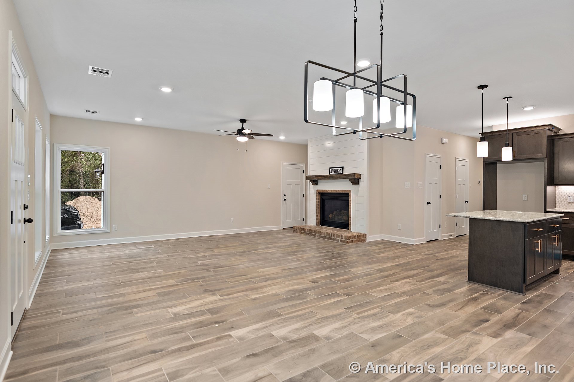 Open floor plan showcasing wood-look tile flooring, shiplap fireplace with brick hearth, large window with decorative trim, modern pendant lights above a stone kitchen island, dark