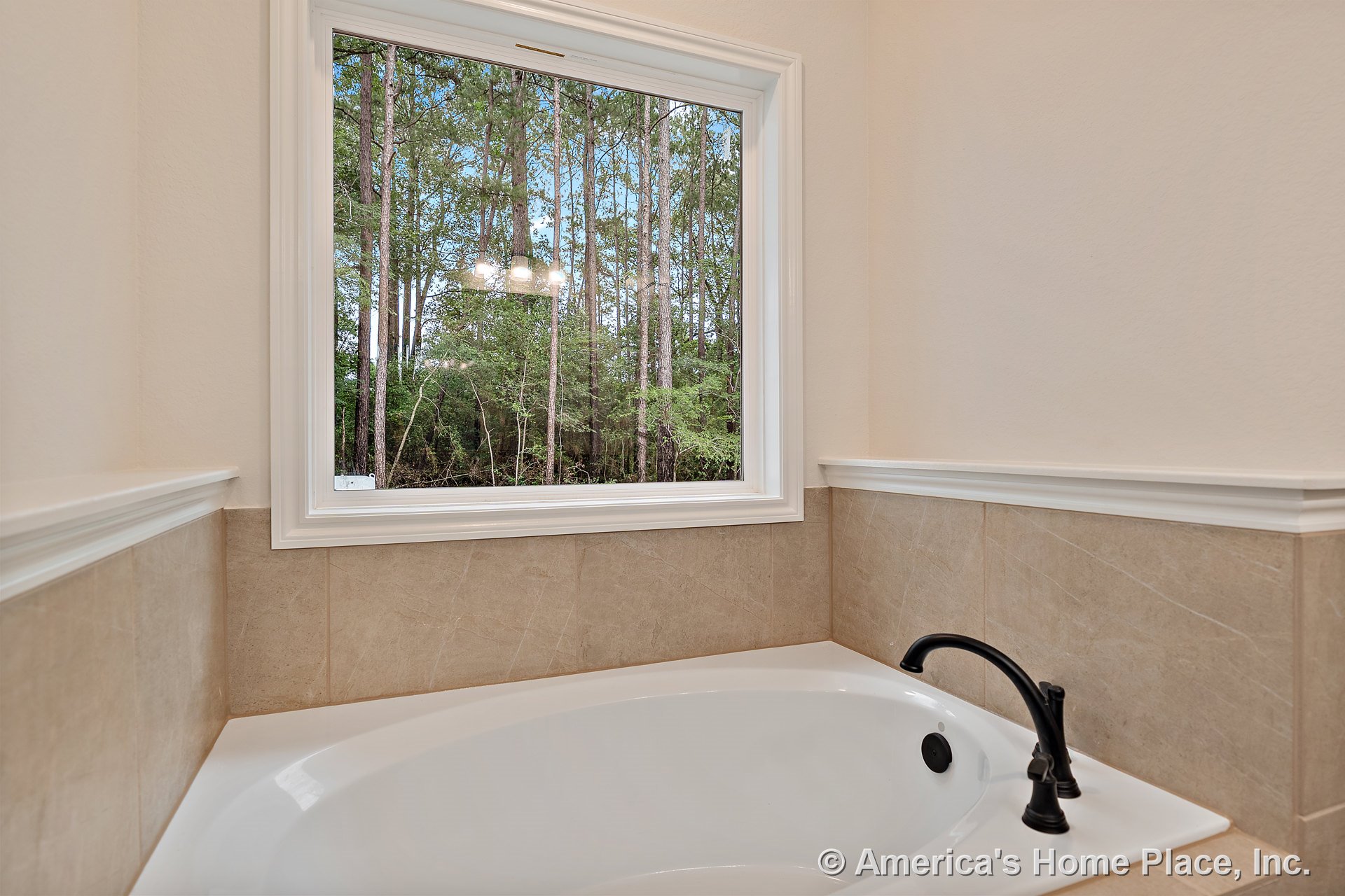 Soaking bathtub with beige tile surround beneath a large picture window framed by white trim and molding, matte black tub faucet, and neutral wall paint.