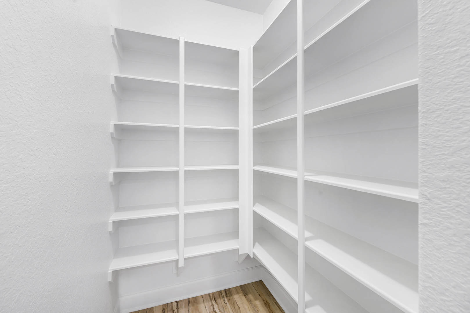 White built-in shelves against a textured white wall, wood flooring with white baseboard, open shelving design in a pantry or storage room.
