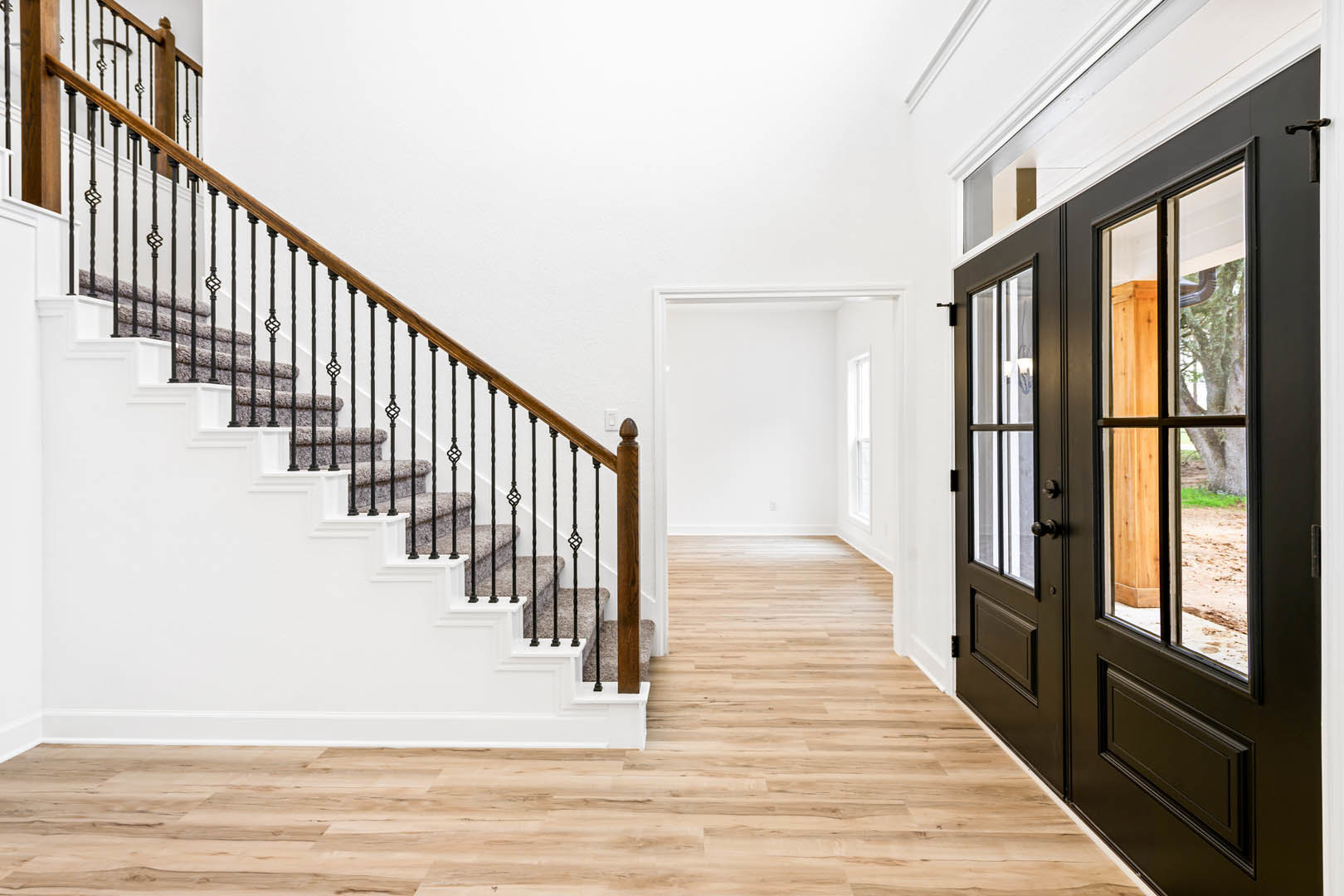 Wood staircase with black metal railing, double glass doors, wide plank wood flooring, white walls, large window in a bright entryway.
