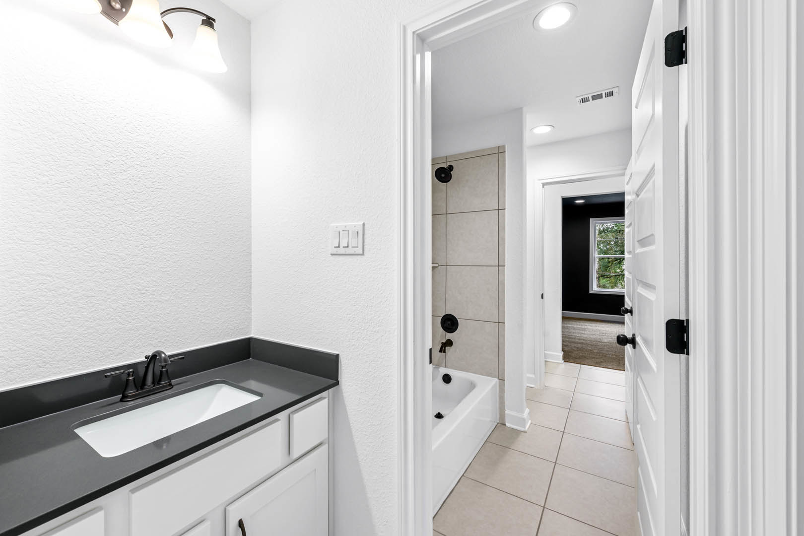 White-framed window above freestanding bathtub, modern sink with chrome faucet on stone countertop, tiled walls, ceiling light, and wall-mounted light switch