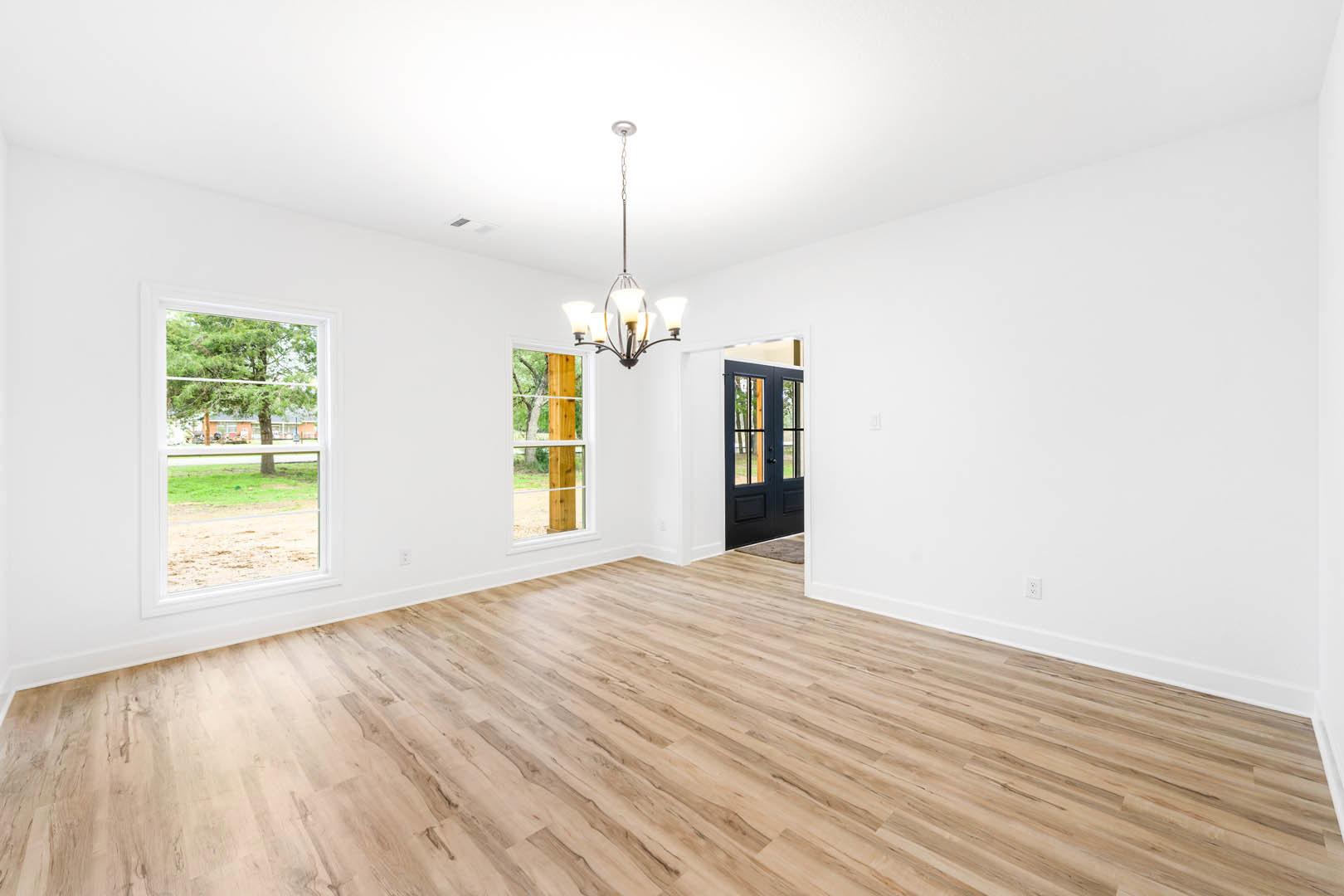 Wood flooring in a spacious room with plaster walls, double doors, a decorative chandelier, and large windows overlooking trees and grass.
