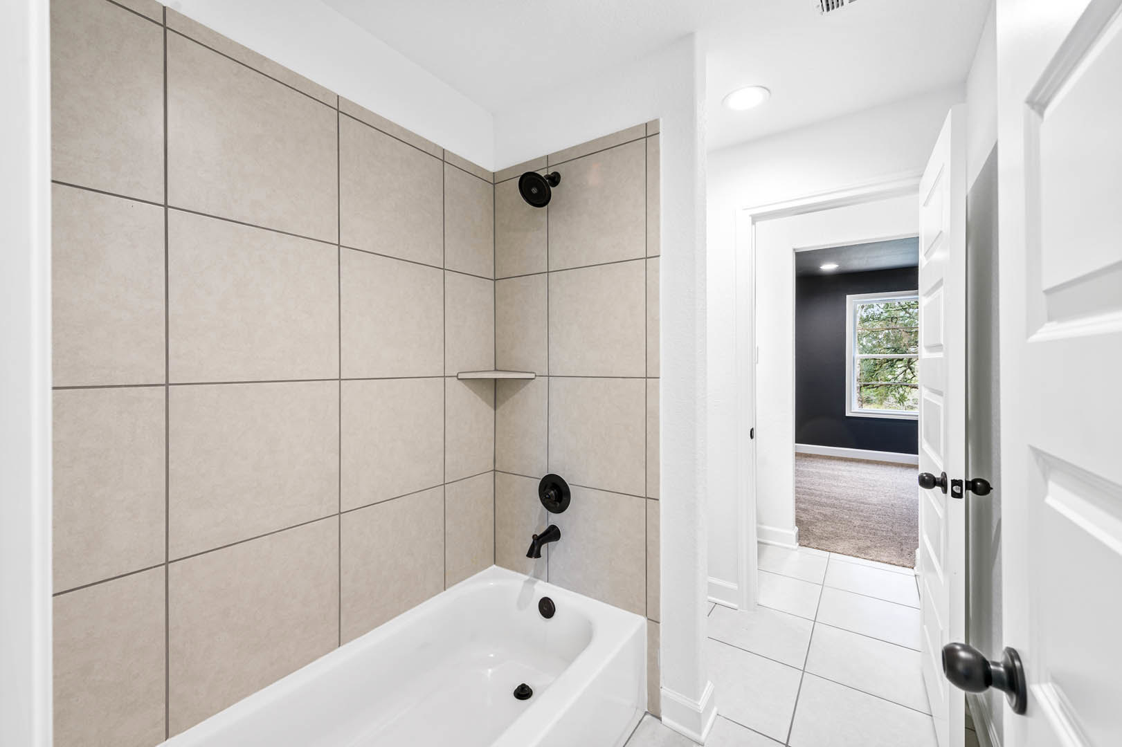 White tile bathroom with freestanding bathtub, matte black faucet, circular black wall fixture, and window with white frame