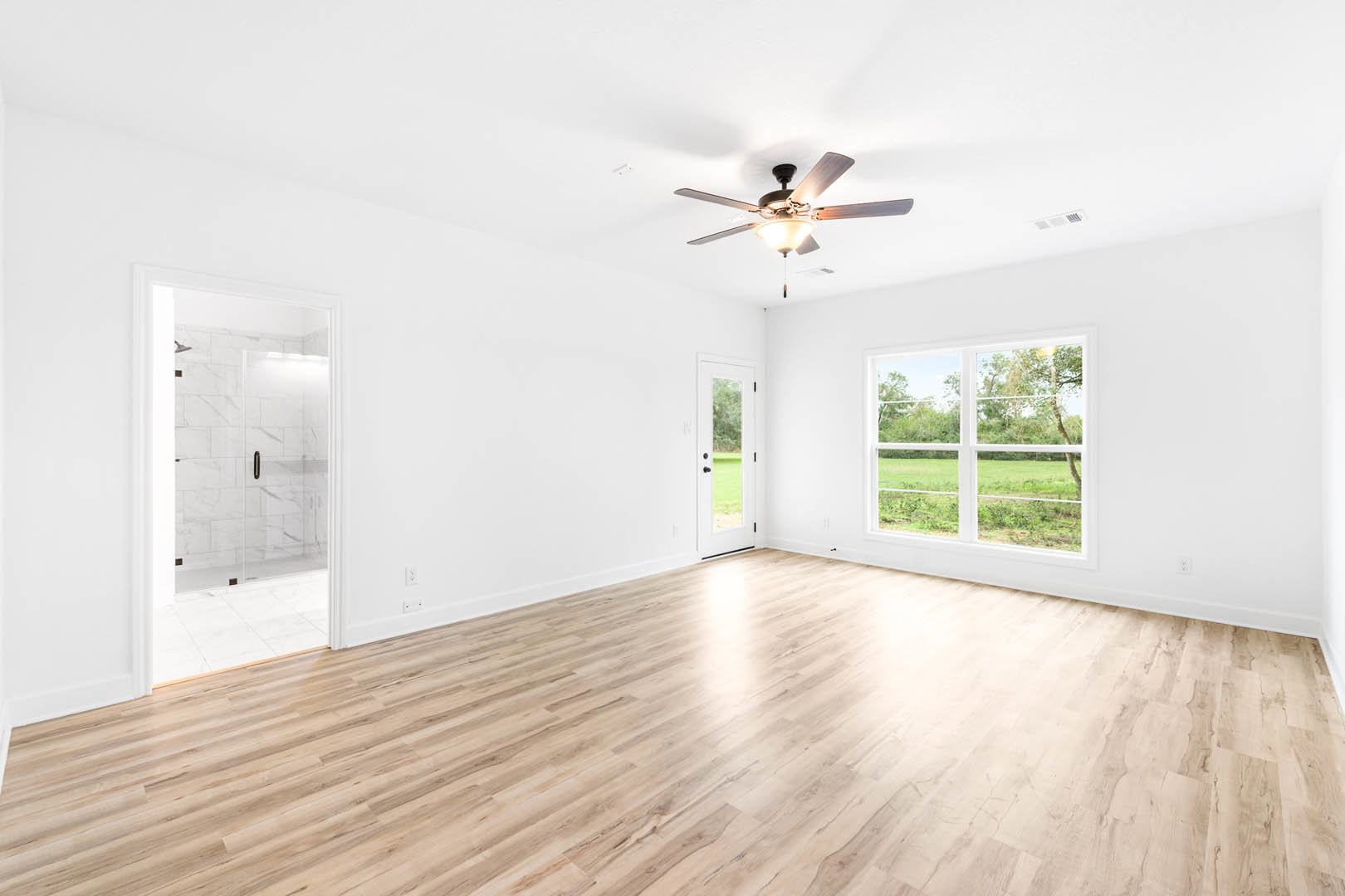 Ceiling fan with light fixture, hardwood flooring, large window overlooking trees, glass shower door with white tile wall, white door with window panel