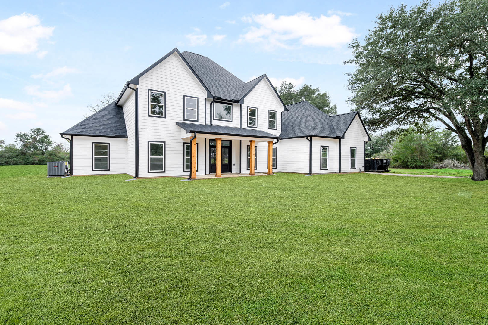 Large white farmhouse with black trim, expansive green lawn, leafy tree, white-framed windows, and cloudy sky.