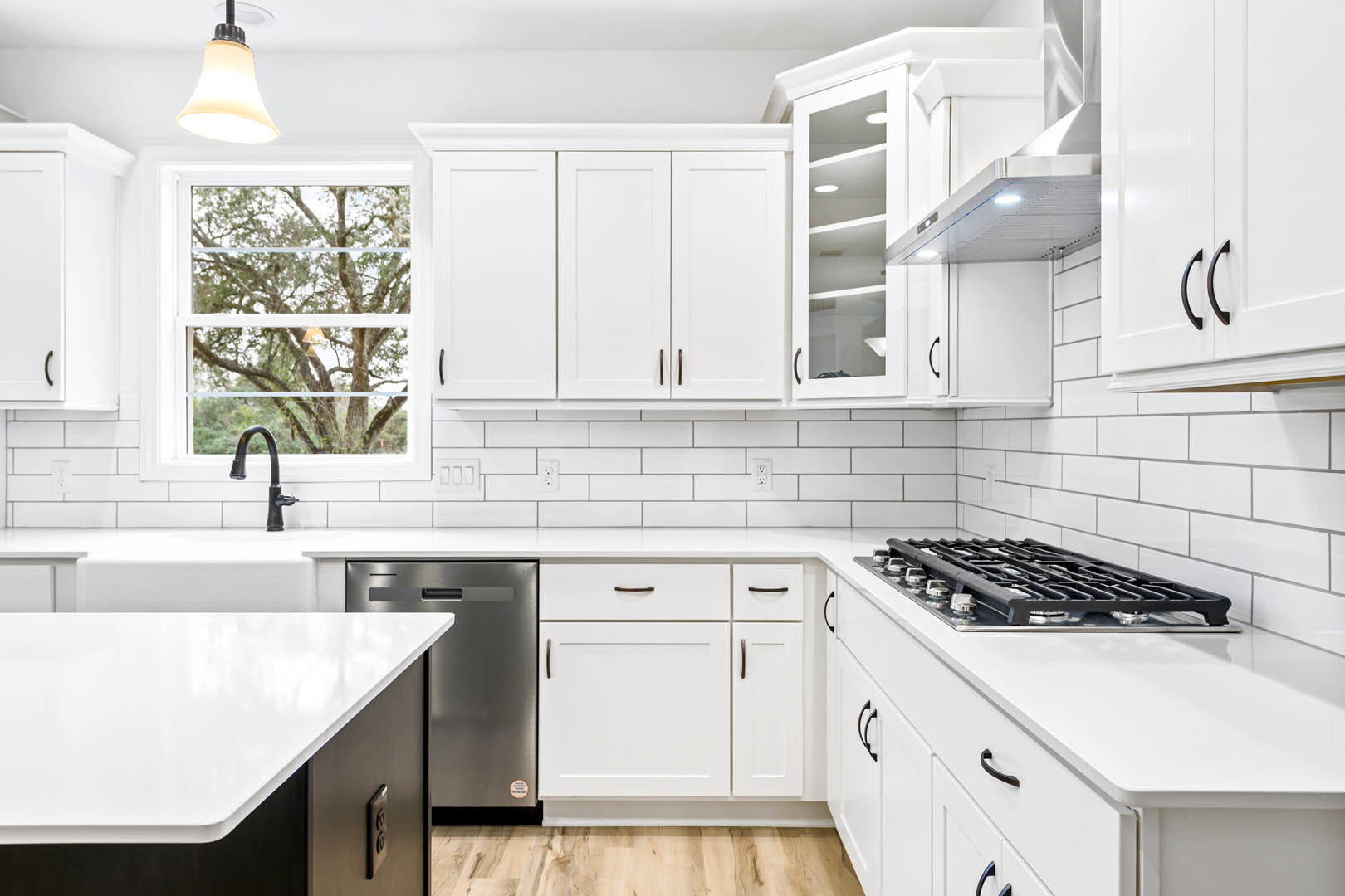 White kitchen cabinets, stainless steel stove, quartz countertops, window overlooking leafy tree, pendant light with white shade, stainless steel refrigerator