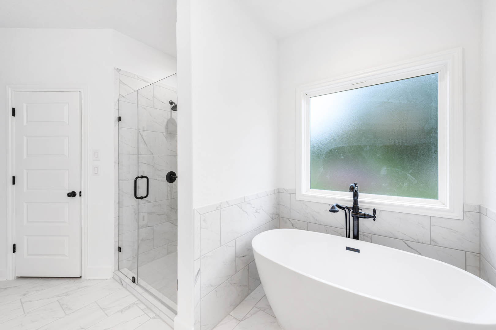 Bathroom with white tub featuring black handle and faucet, glass shower enclosure, frosted window, white tile walls, and white door with black hardware.