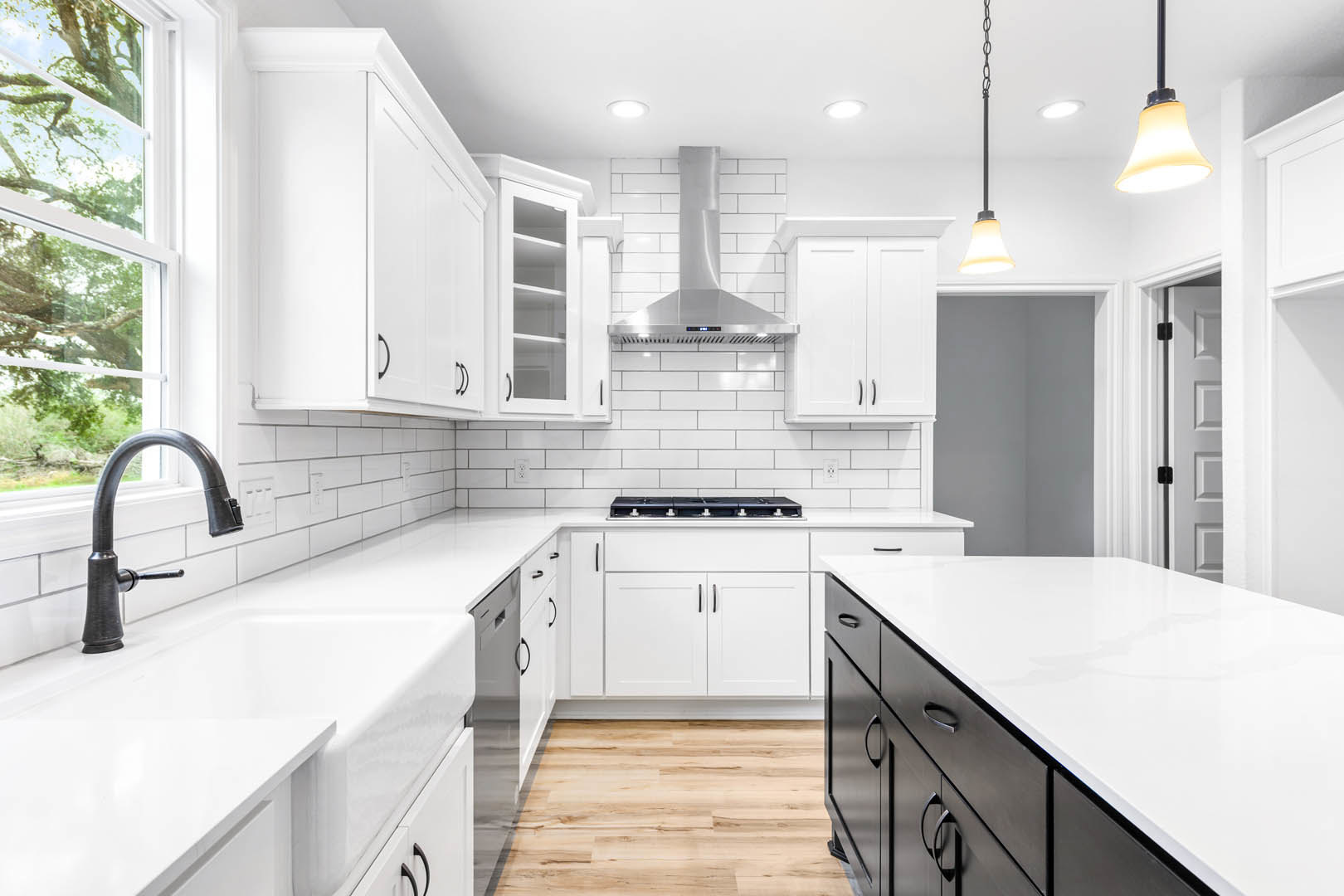 White kitchen cabinets and black drawers beneath a white countertop, stainless steel stove with matching exhaust hood, black faucet at the sink, wood flooring with white trim