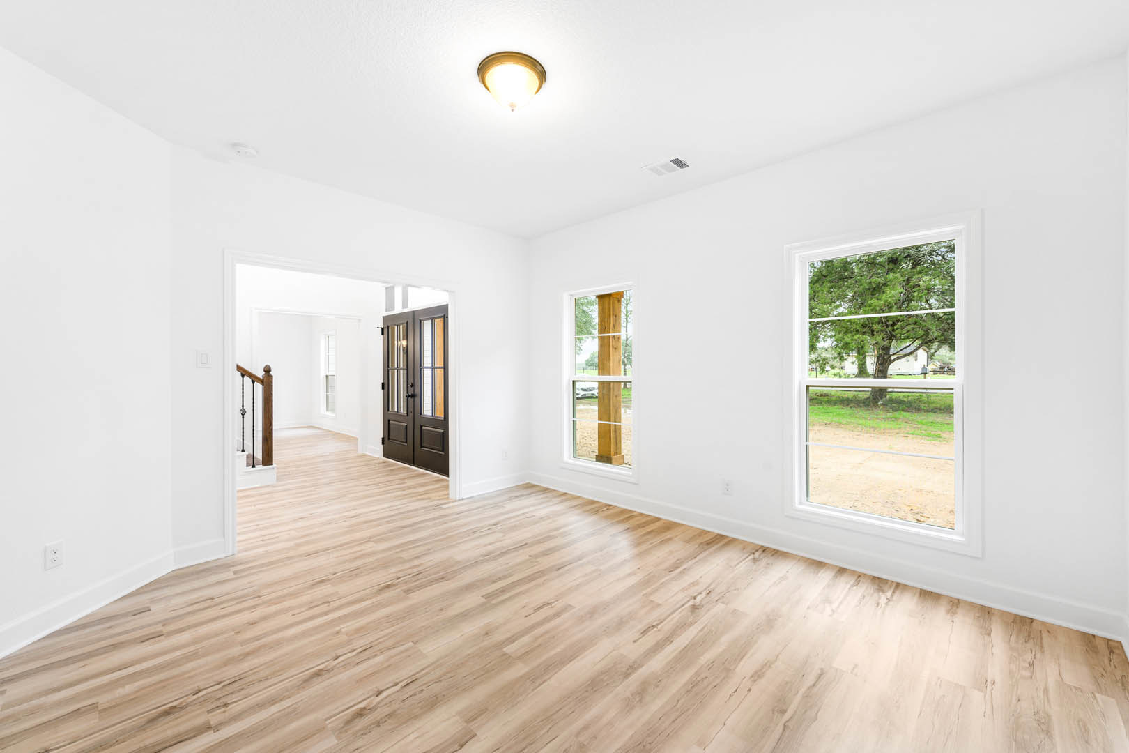 Spacious room with light wood flooring, large windows overlooking trees, white plaster walls, modern ceiling light fixture, and a close-up view of a wooden door