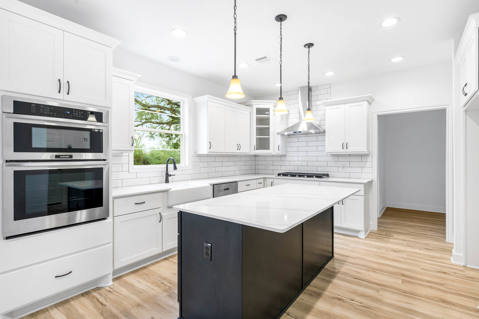 White shaker cabinets and black island with white countertop, stainless double oven and microwave, chrome faucet, window overlooking tree, light wood flooring