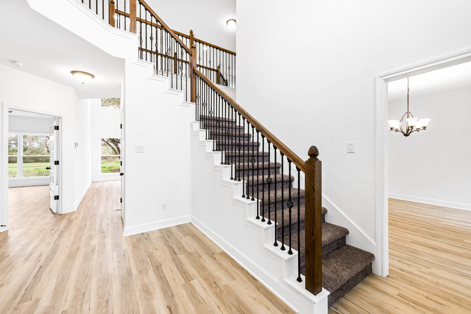 Carpeted staircase with metal and wood railings, white walls, open white door revealing grassy field, illuminated ceiling light fixture, laminate and wood flooring