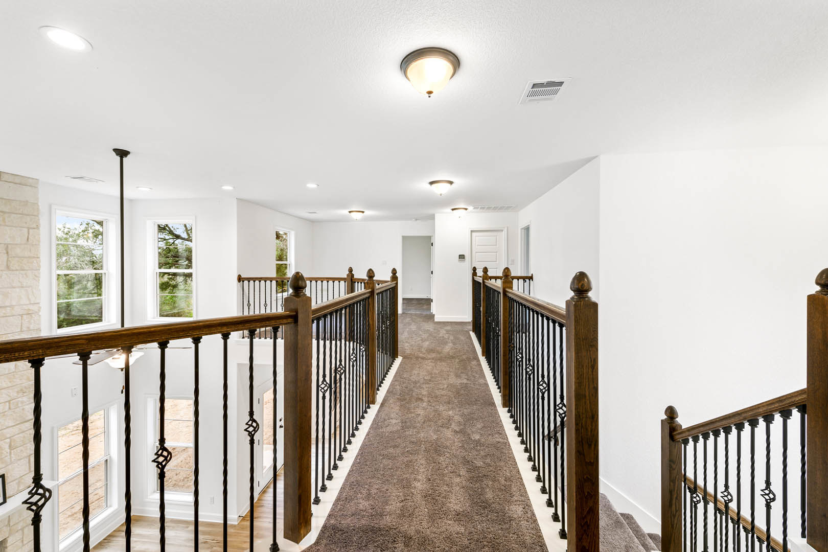 Hallway with brown carpet, wood and metal railing along staircase, ceiling vent, light fixture, and window overlooking trees