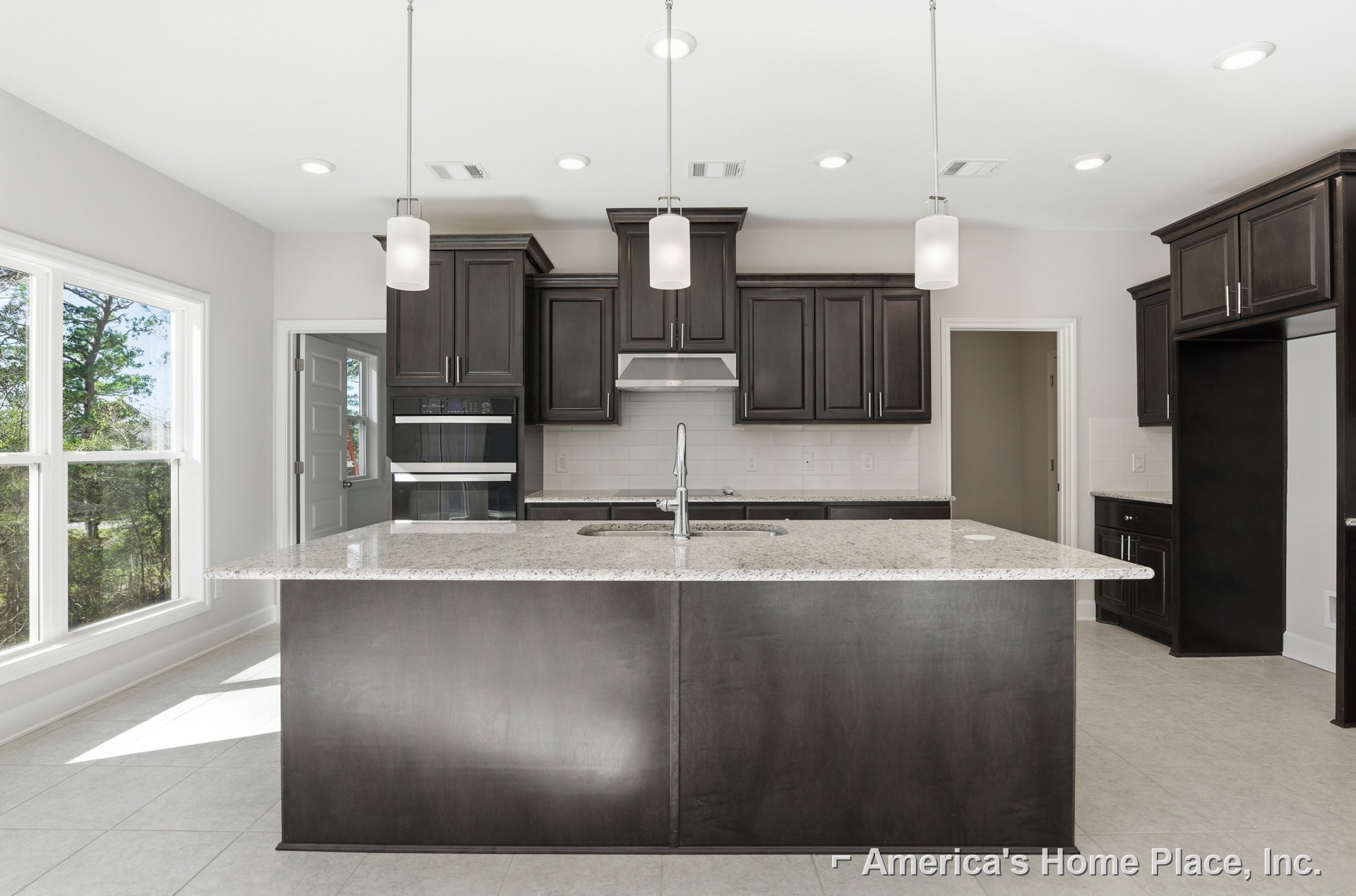 Spacious kitchen featuring a large central island with white countertops, black oven with glass doors, white cabinetry, stainless steel sink, and a window overlooking green trees.