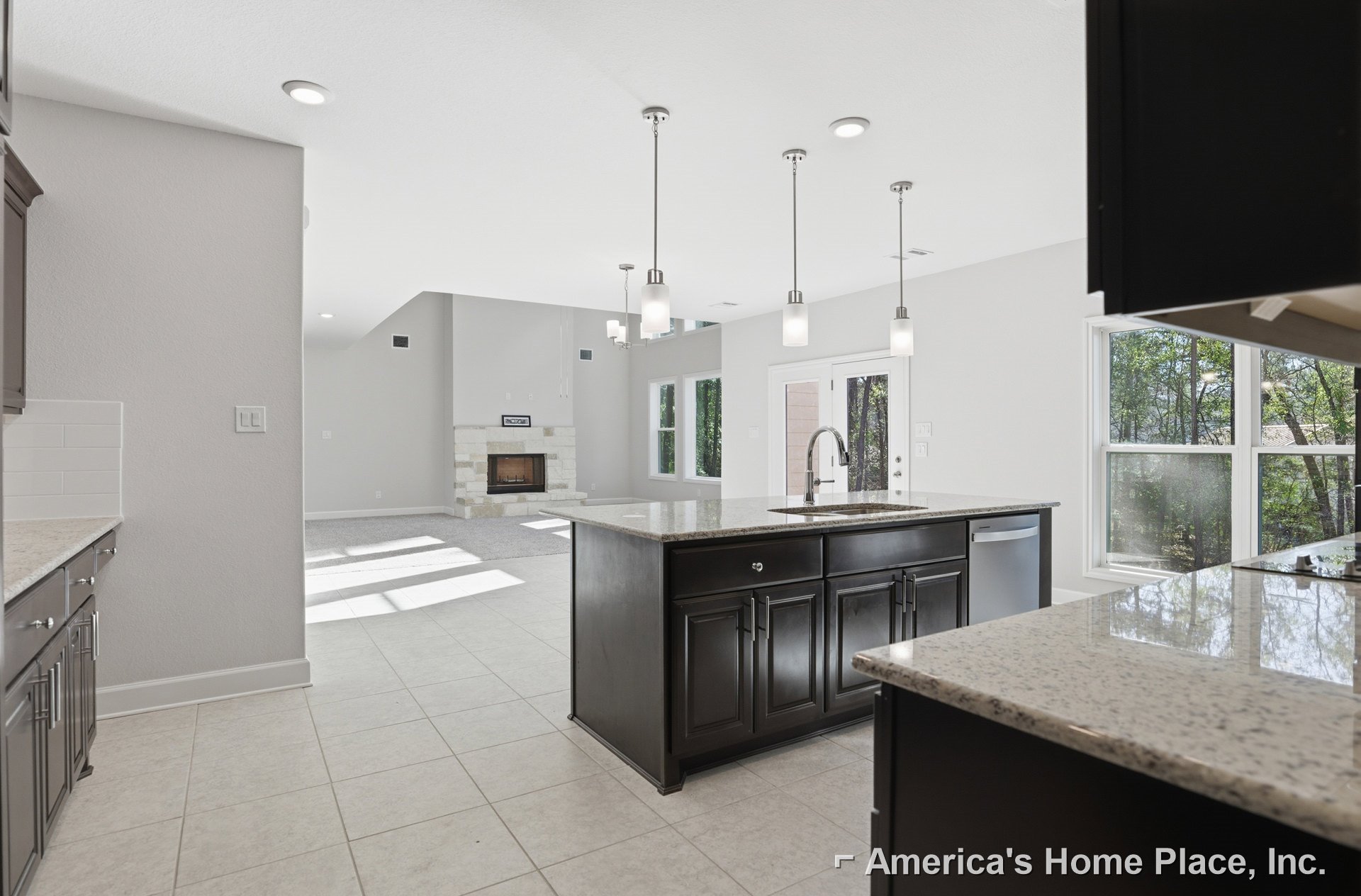 Spacious kitchen featuring a large central island with white granite countertops, tile flooring, wood cabinetry, stainless steel sink and faucet, and a fireplace with a wood