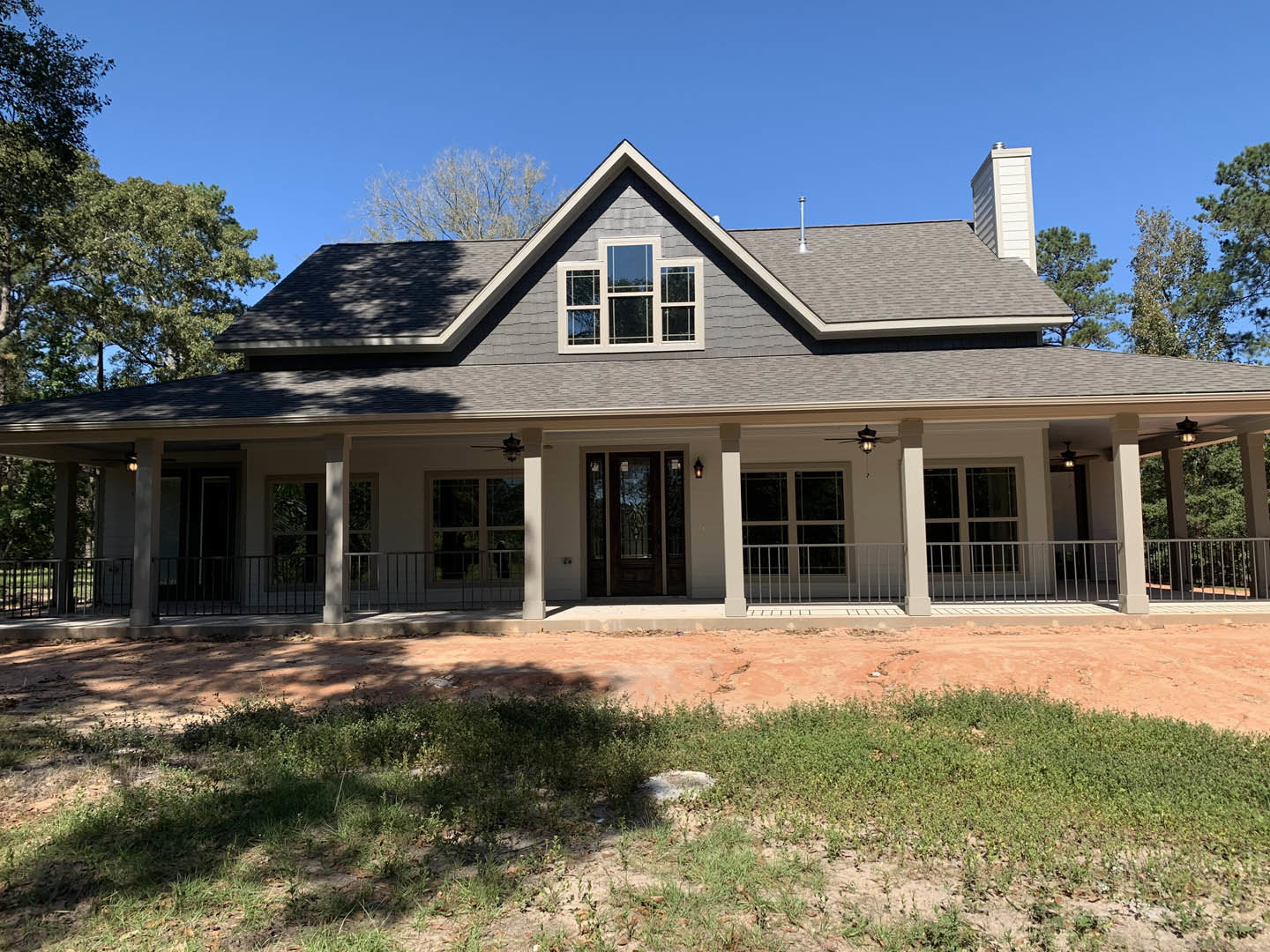 White house with covered porch, wooden railing, and picket fence; large windows reflect blue sky; dirt patch with young plants in front; Sidney Lanier Cottage visible in