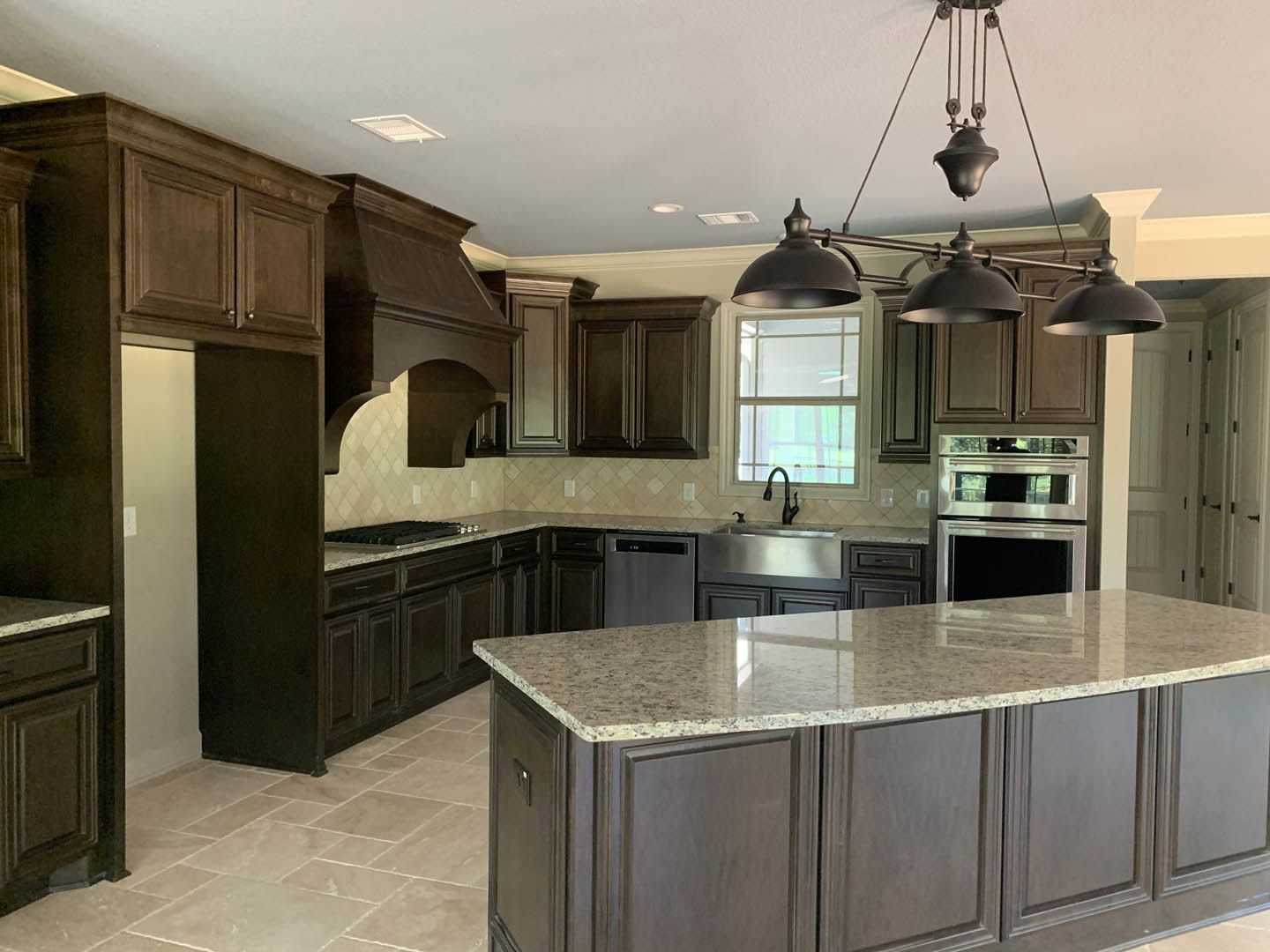 Spacious kitchen featuring a large central island with stone countertop, white cabinetry, stainless steel double oven, farmhouse sink beneath a window, and pendant light fixtures