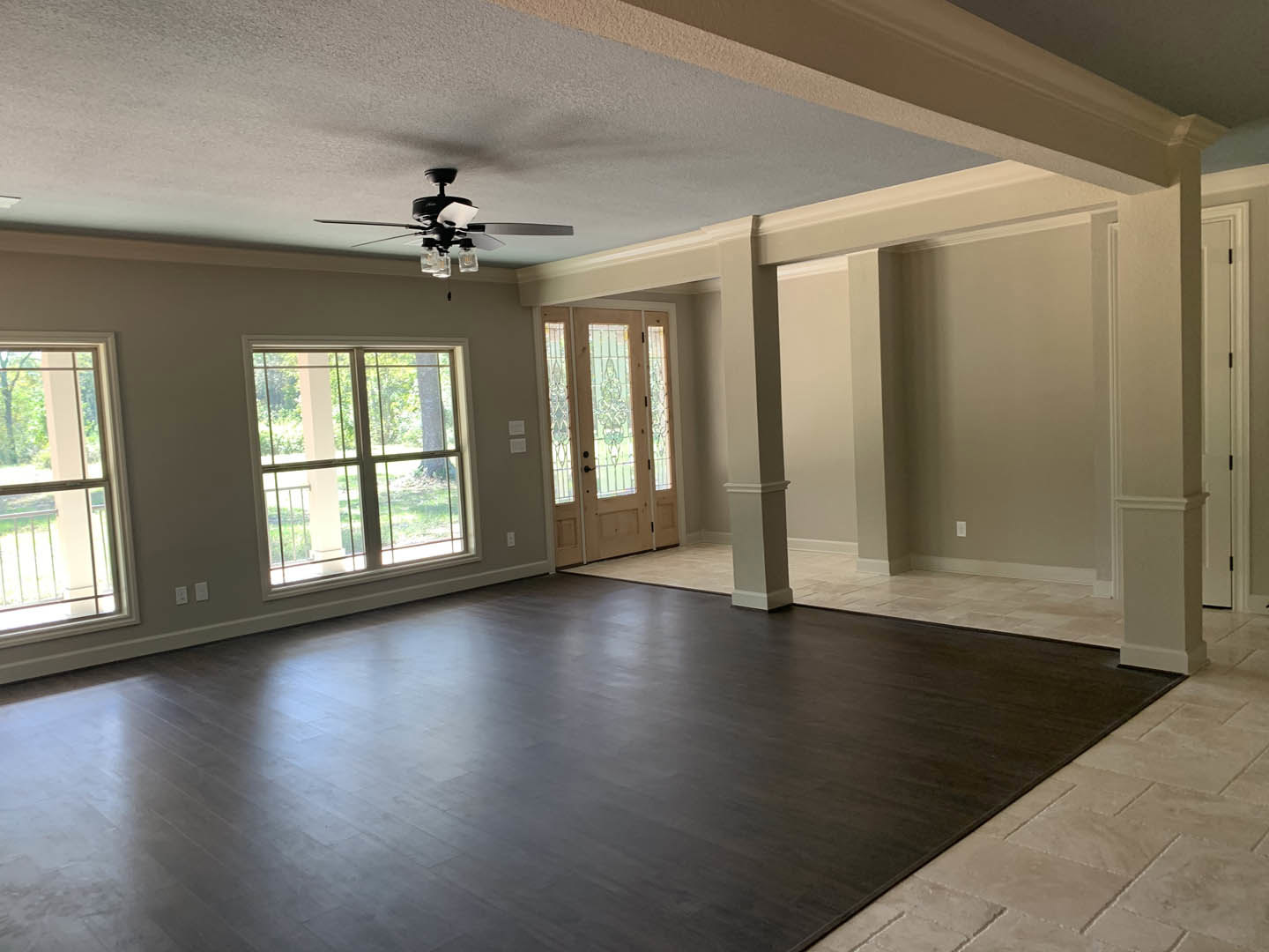 Spacious living area with wood laminate flooring, ceiling fan with lights, large window overlooking trees and railing, glass-paneled door, white plaster walls and ceiling