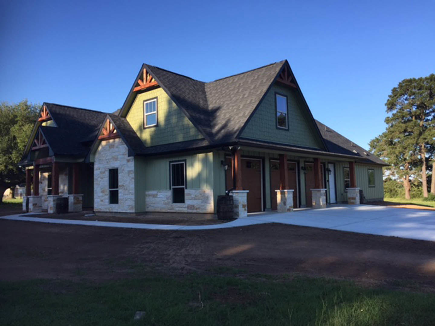 Two-story home with gray shingle roof, white-trimmed windows, paved driveway, and mature tree with green leaves in front yard under blue sky