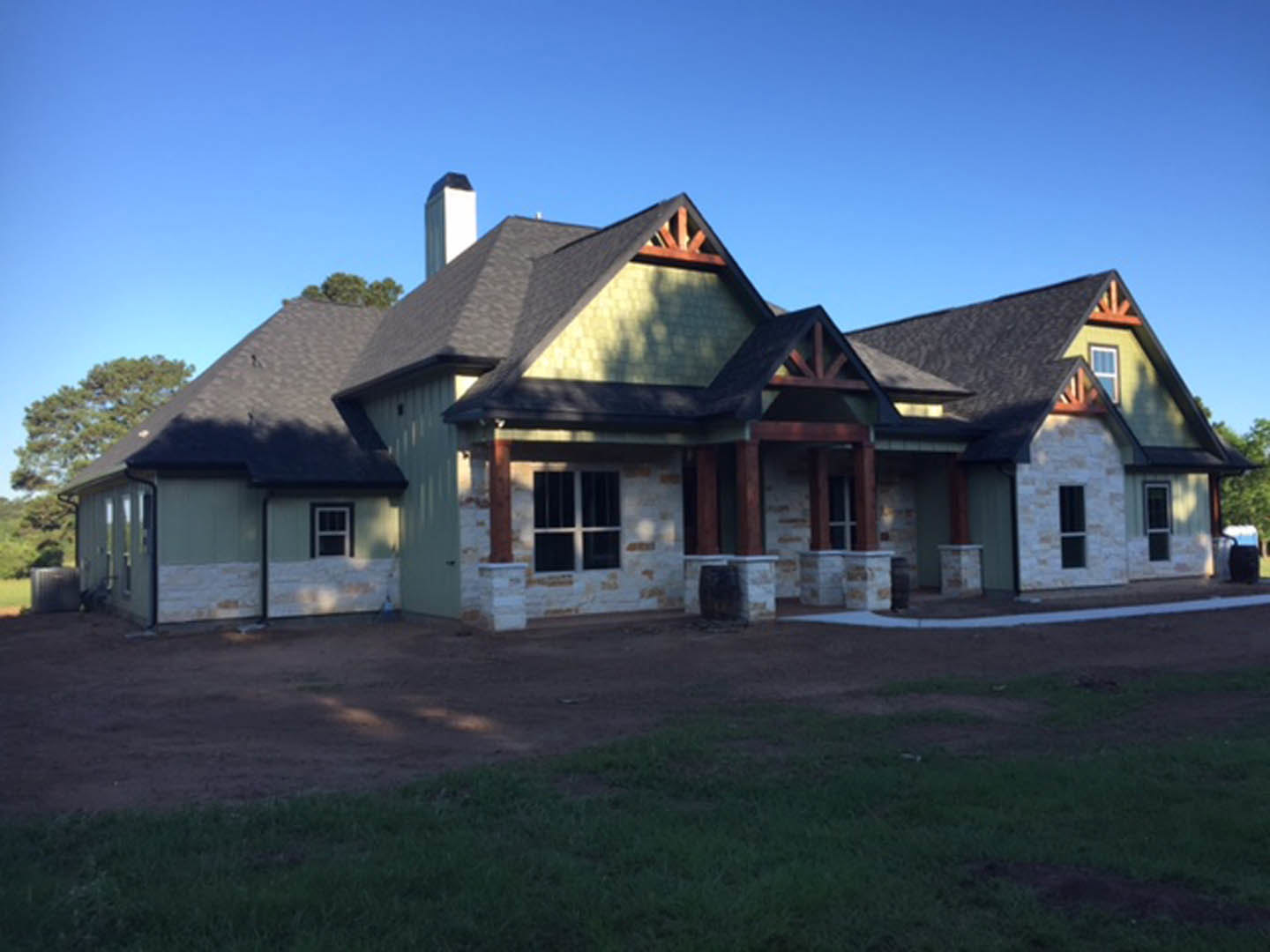 Green metal roof atop a cottage-style home with white-framed windows, brick chimney, covered porch, and grassy lawn bordered by trees.