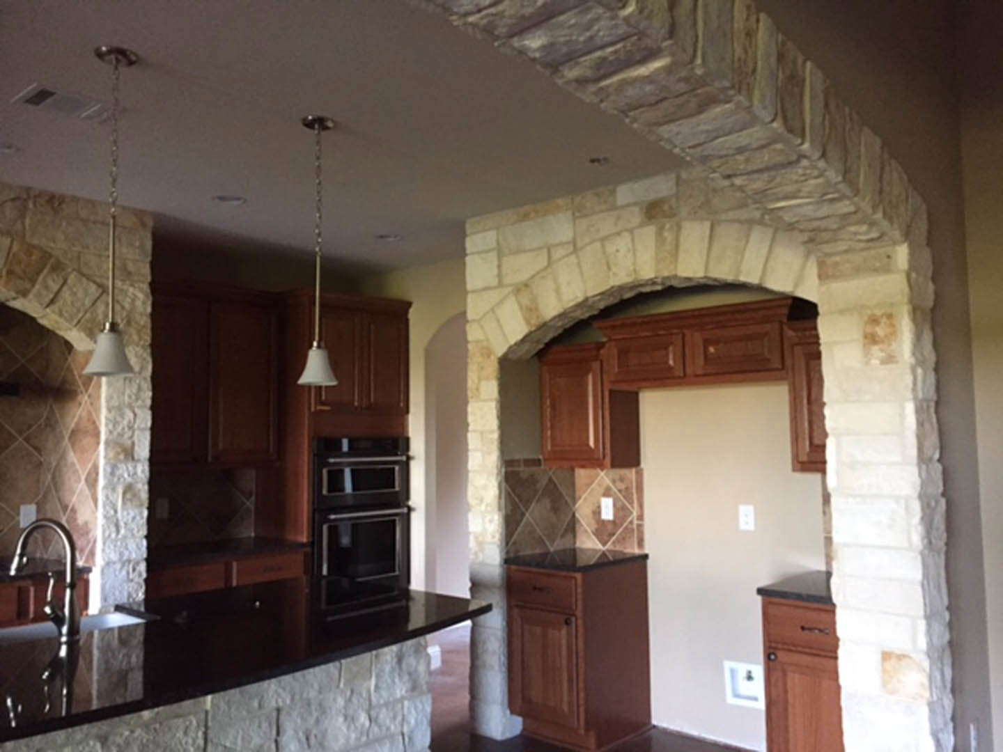 Kitchen with textured white stone walls, dark wood cabinets, black oven, black countertop, hanging chain from ceiling, sink, and drawers