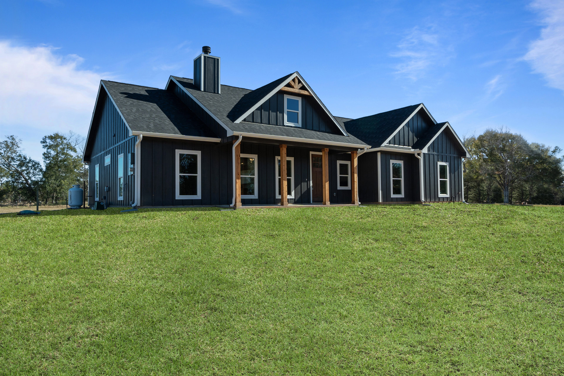 White farmhouse with large windows, front door with glass panel, brick chimney, and green lawn bordered by landscaping under a partly cloudy sky