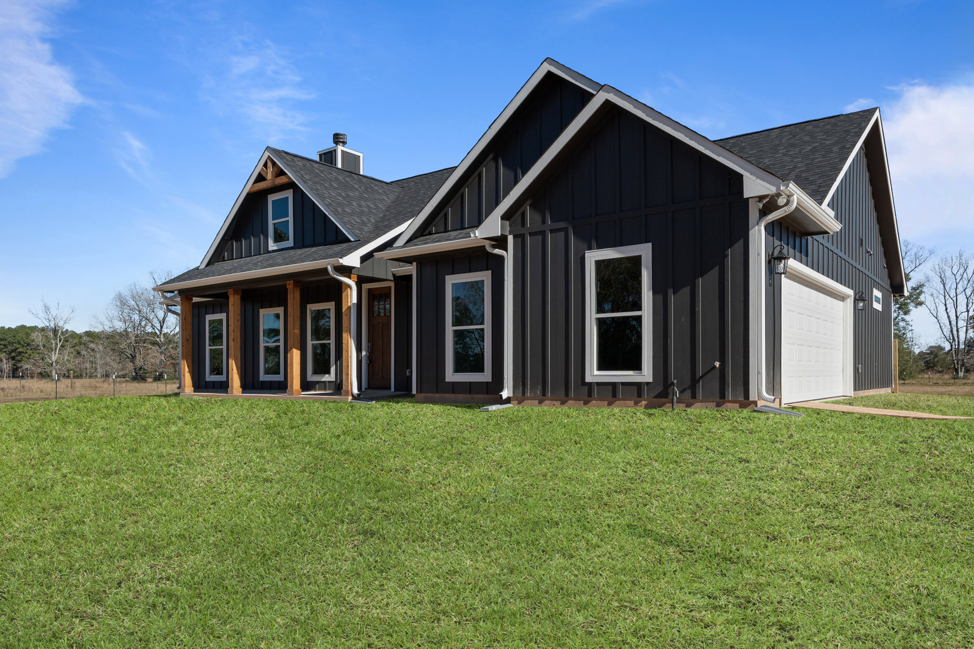 Two-story house with white siding, black-framed windows, white garage door, and green grass lawn; front door and windows reflect surrounding trees.