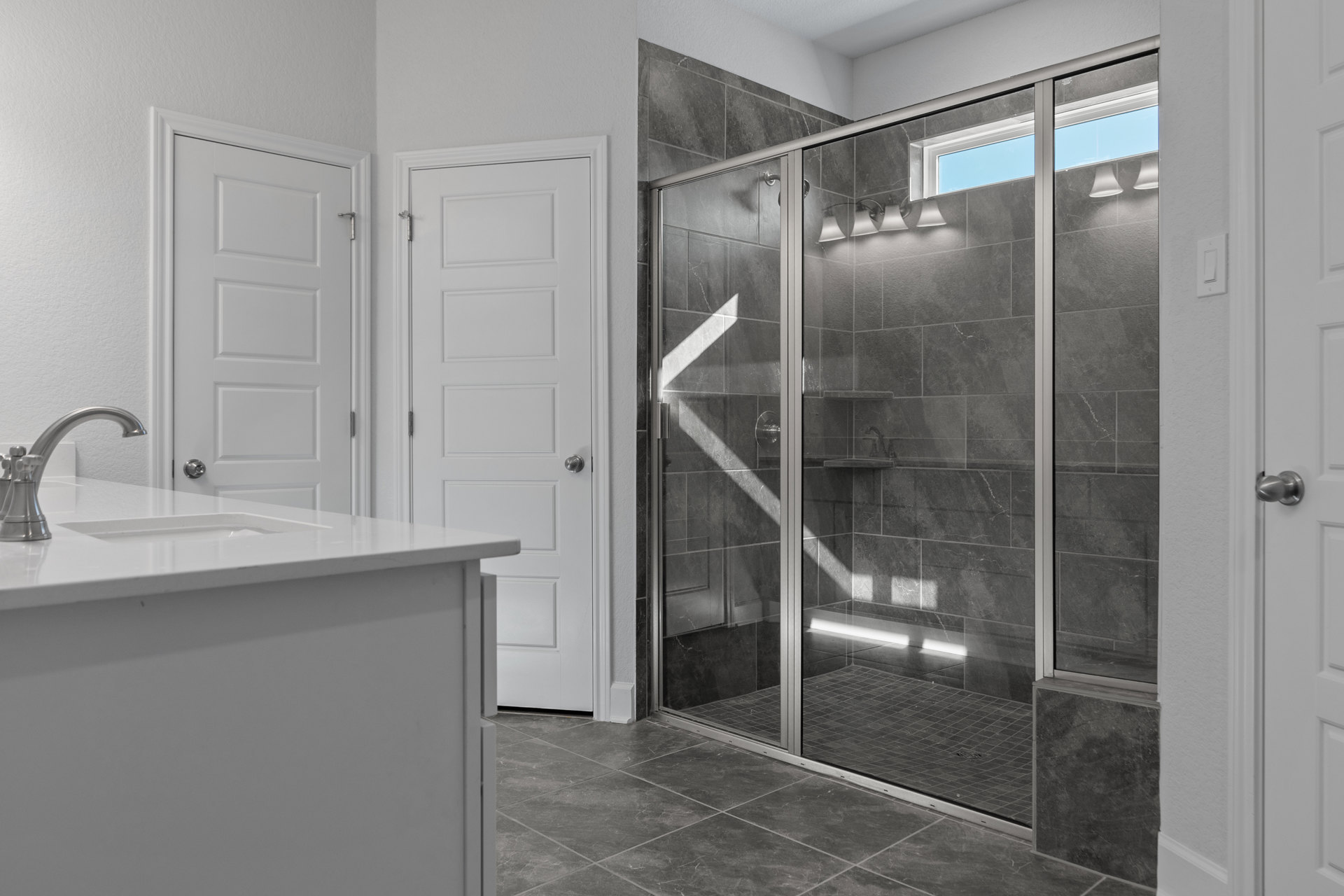Modern bathroom featuring a glass shower enclosure, white countertop with silver faucet, white cabinetry with silver handles, and a white door with a silver knob.