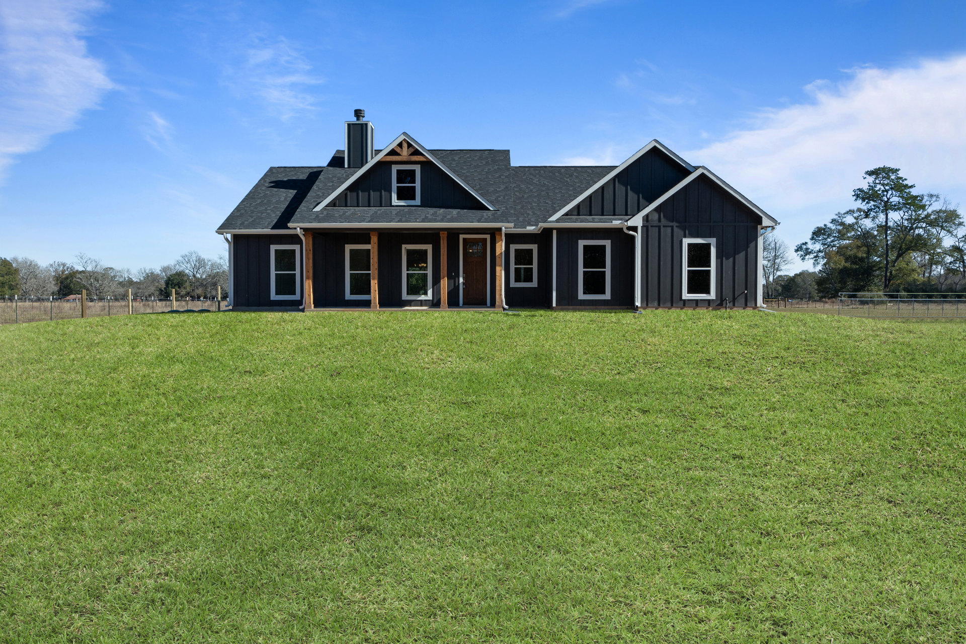 Black farmhouse-style home with white-framed windows, prominent roof and chimney, set behind a spacious green lawn and scattered trees under a cloudy sky